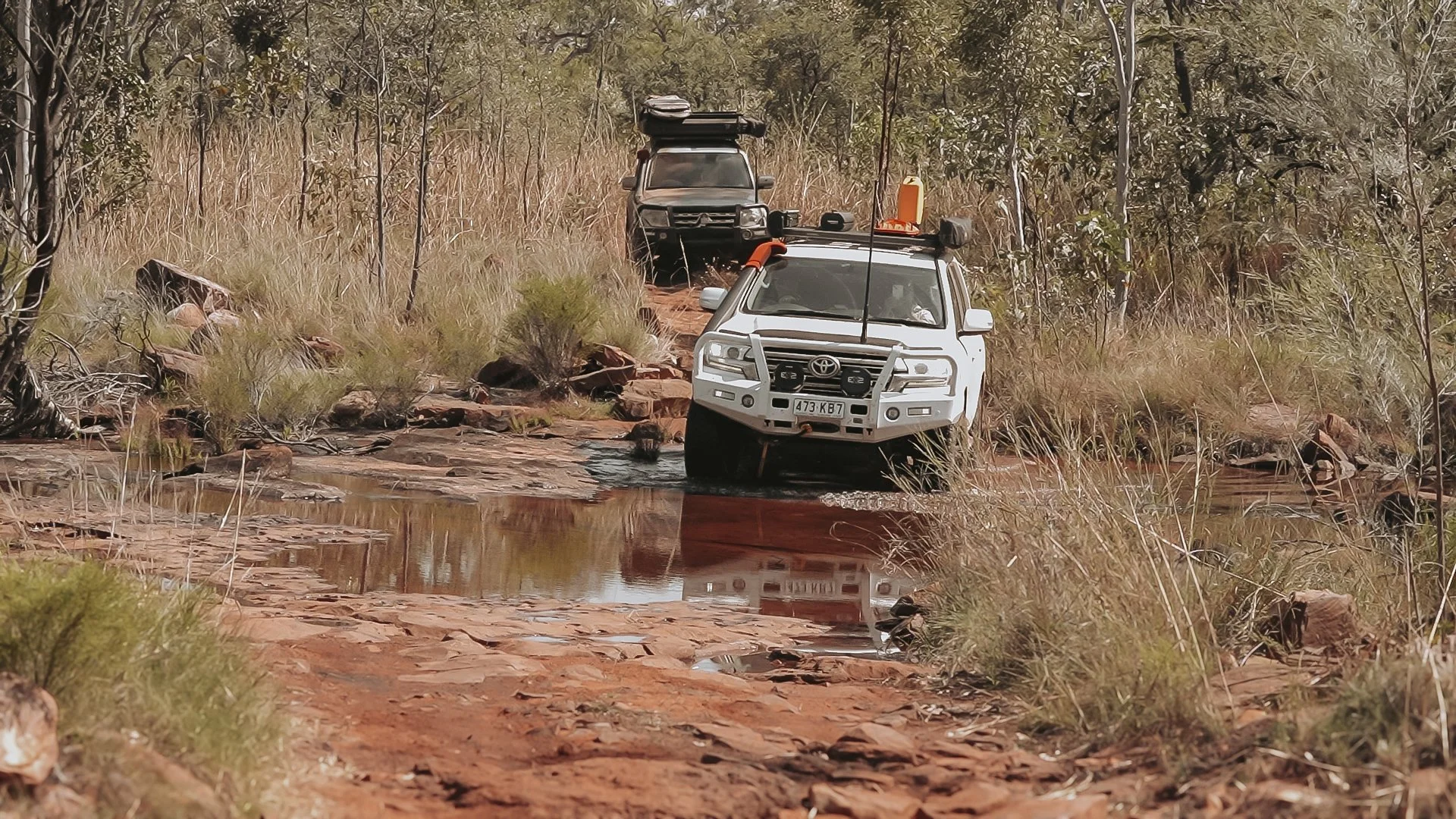 Two off-road vehicles driving through a shallow water crossing on a dirt trail surrounded by dry grass and sparse trees.