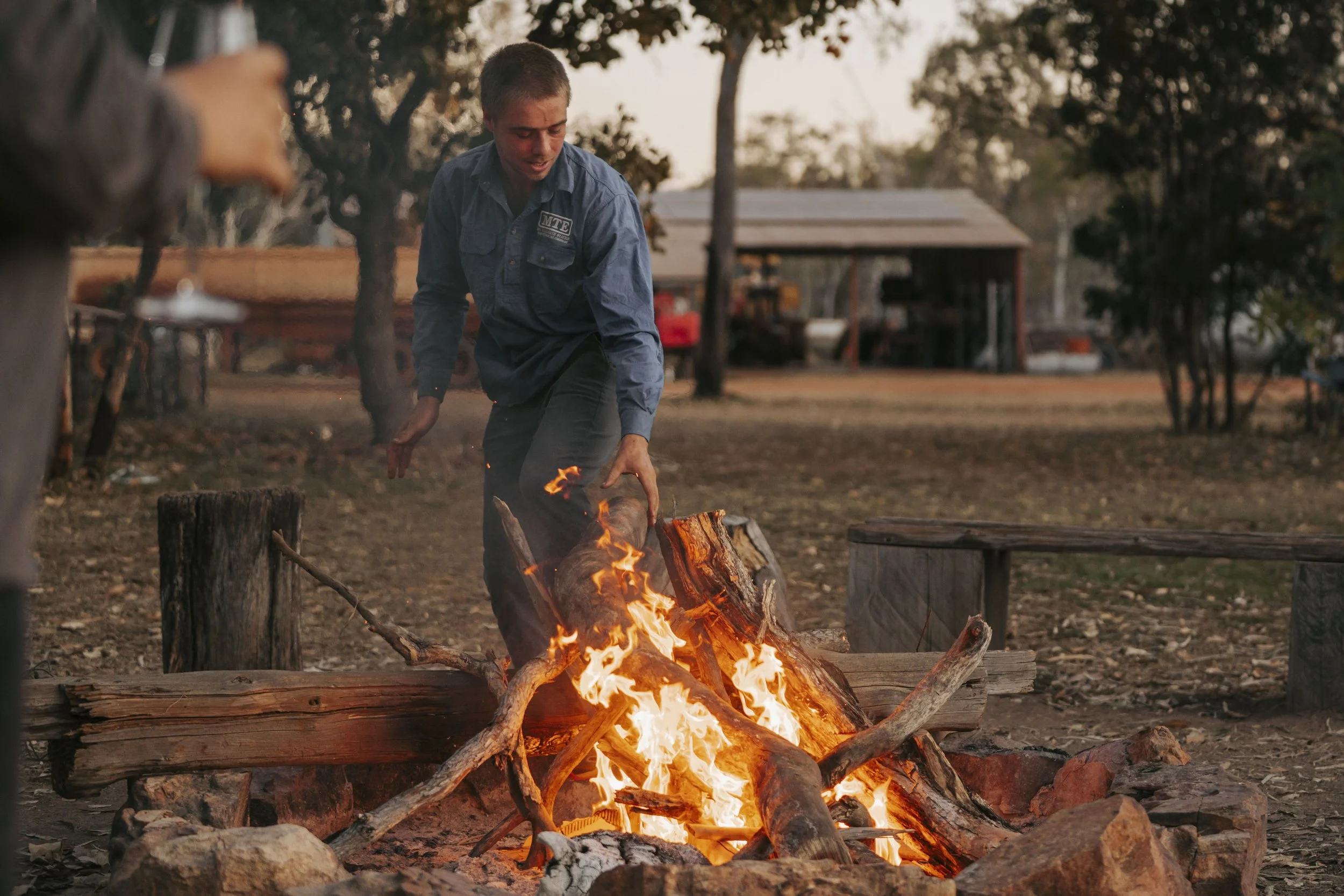 A person inside a fire pit with a wood fire, reaching down to adjust or add more wood, in an outdoor setting during sunset.