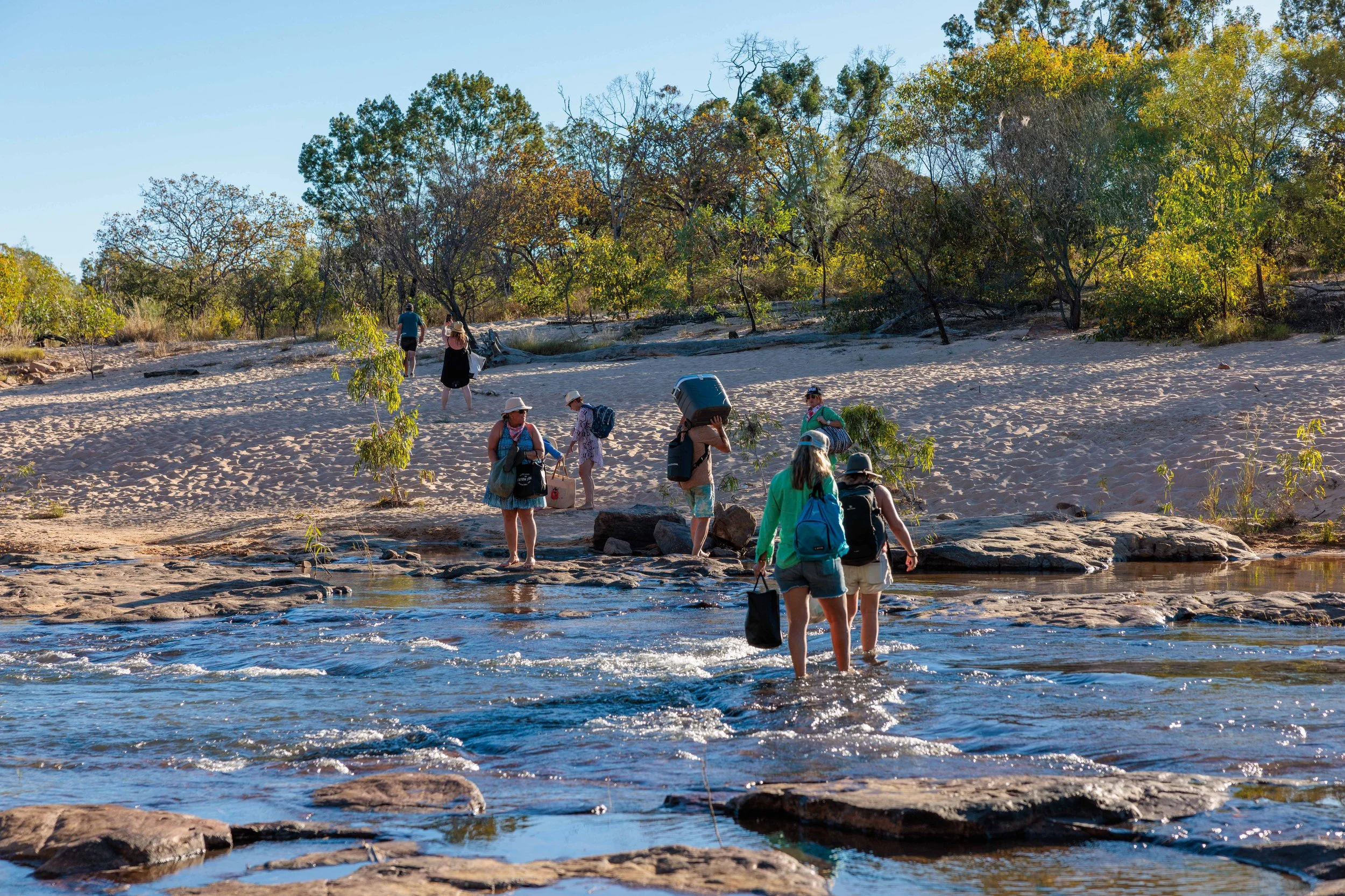 People crossing a shallow river with rocks on a sandy shore, carrying backpacks and gear, surrounded by trees with green and yellow leaves under a clear blue sky.