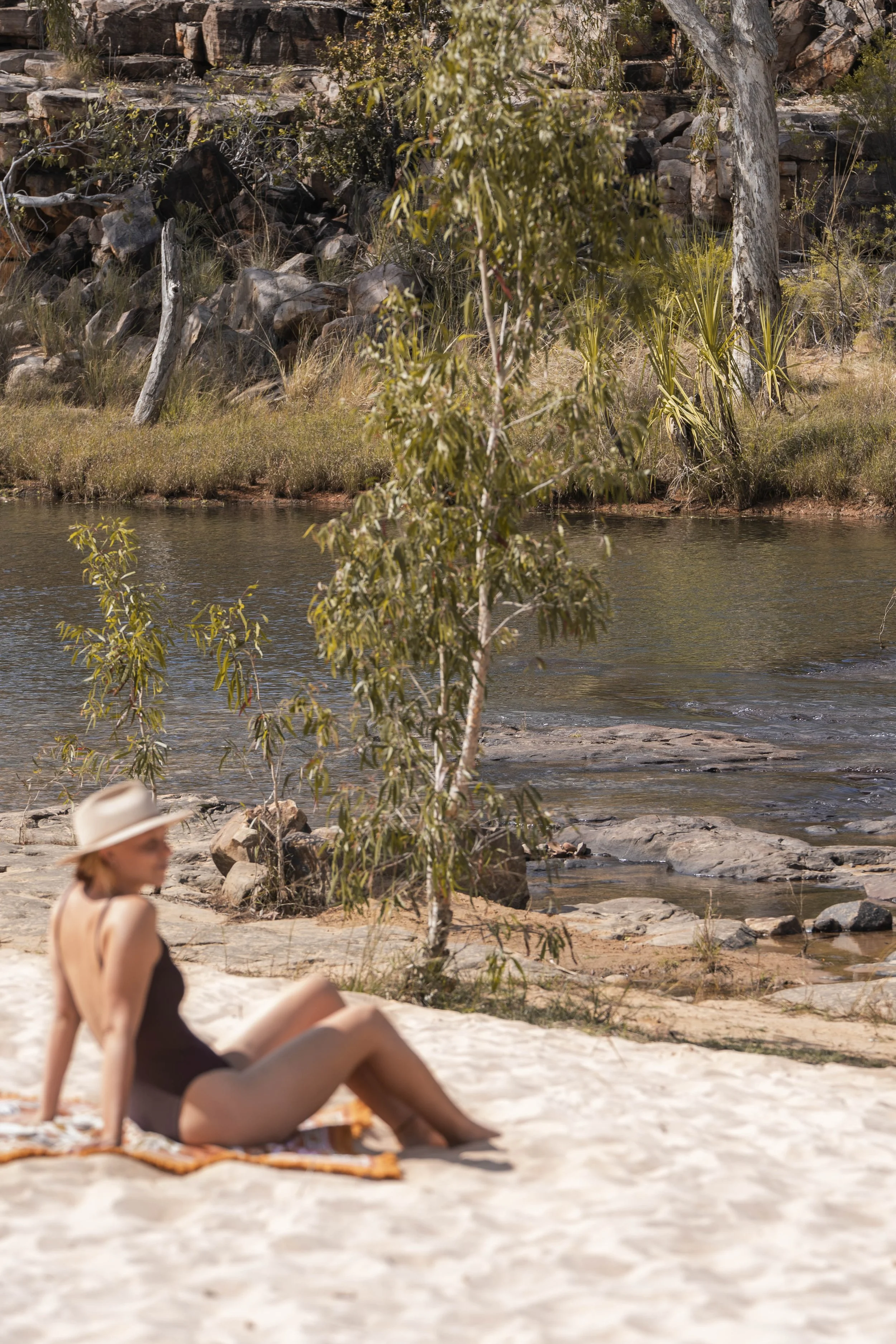 A woman in a black swimsuit and a wide-brimmed hat relaxing on a towel by a river in a natural outdoor setting with trees and rocks.