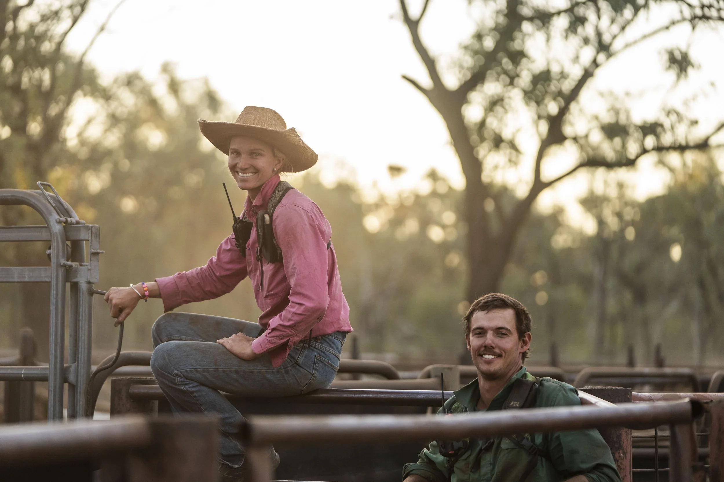 A woman in a pink shirt and a straw hat riding a horseback with a man in a green shirt, both smiling outdoors with trees in the background