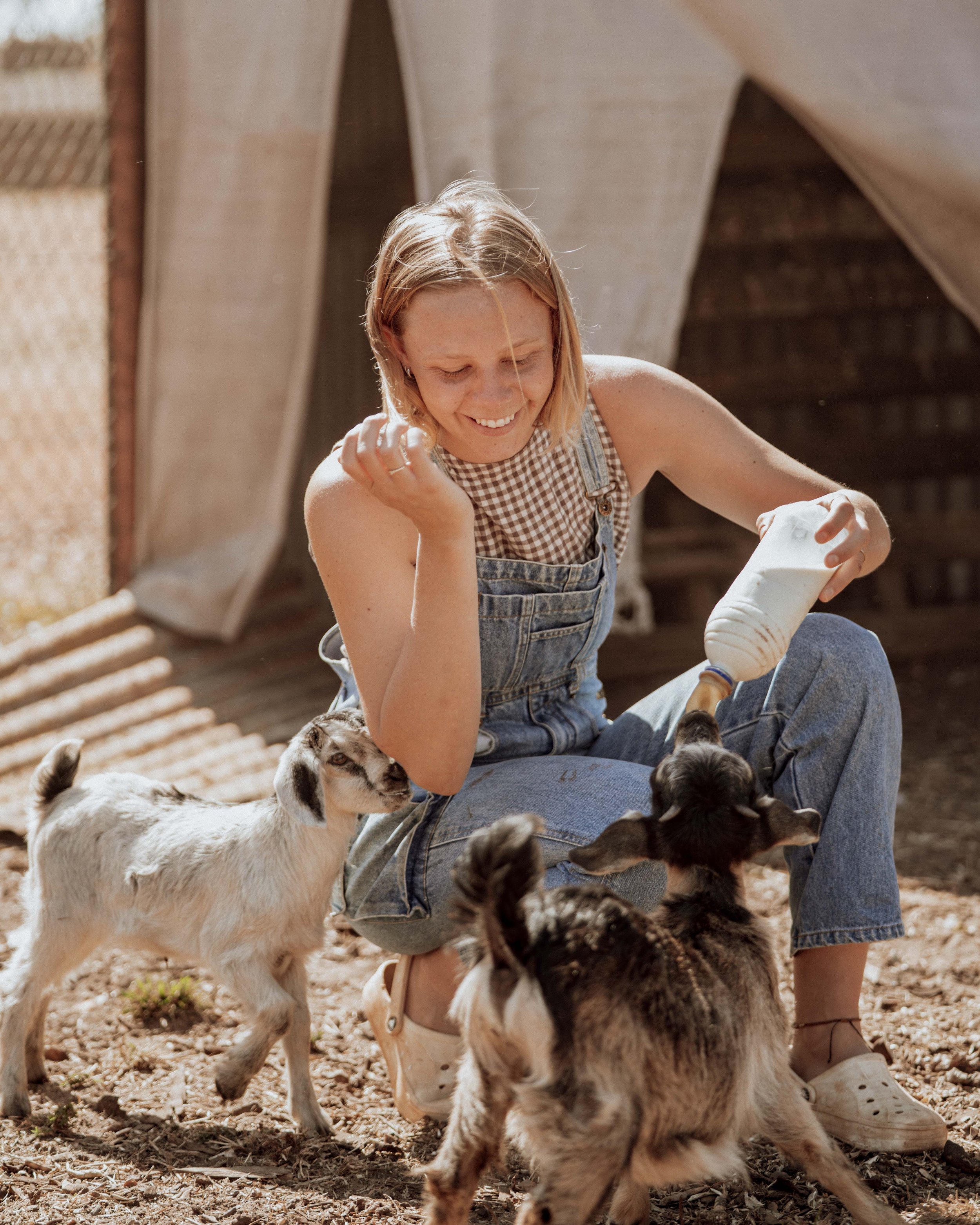 A smiling woman in overalls feeding baby goats on at the Mt Elizabeth Homestead.