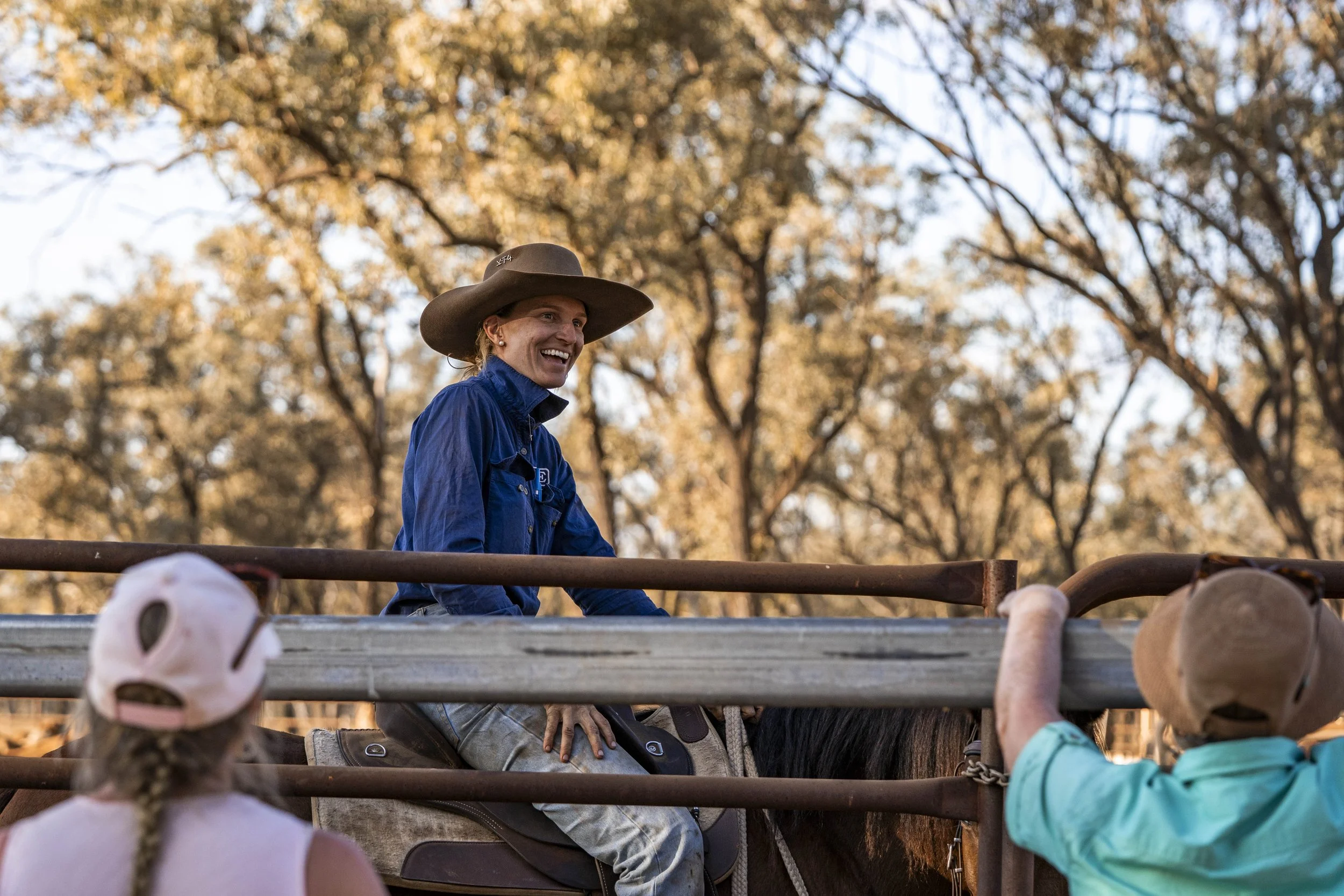 A woman riding a horse, smiling, with two people on the ground talking to her, during sunset with trees in the background.