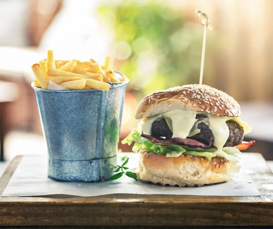 A Burton Beef burger with melted cheese, lettuce, tomato, and beef patty on a bun, served with a side of chips in a small metal bucket on a wooden board.