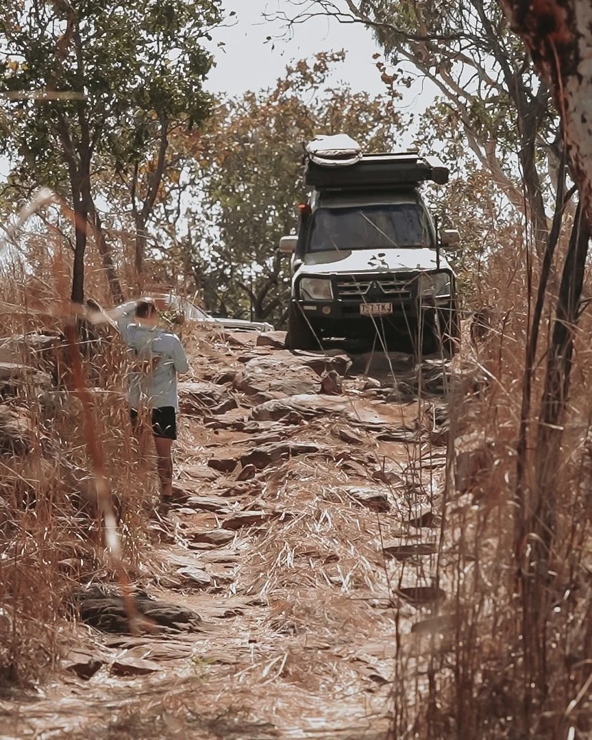 A person walking on a rocky dirt trail with dry grass on both sides, leading to a parked off-road vehicle among trees on the road to Wunnamurra Gorge, an epic 4wd track.