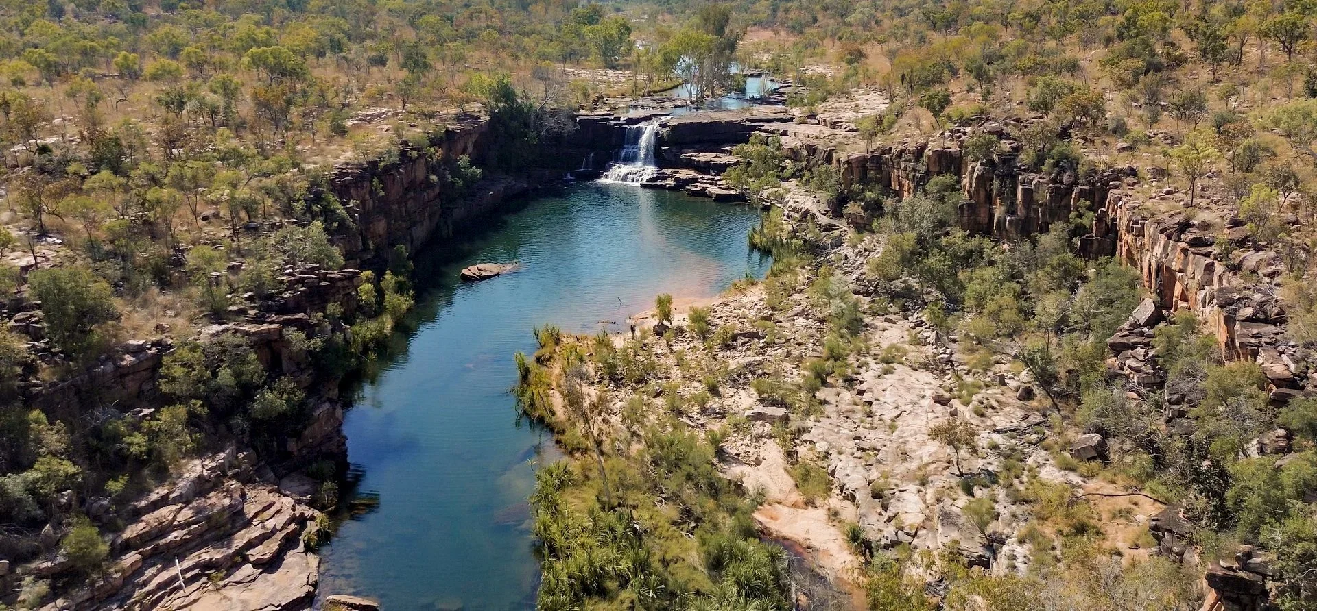 Aerial view of a river in a canyon with a small waterfall, surrounded by rocky cliffs and sparse green vegetation under a clear sky.