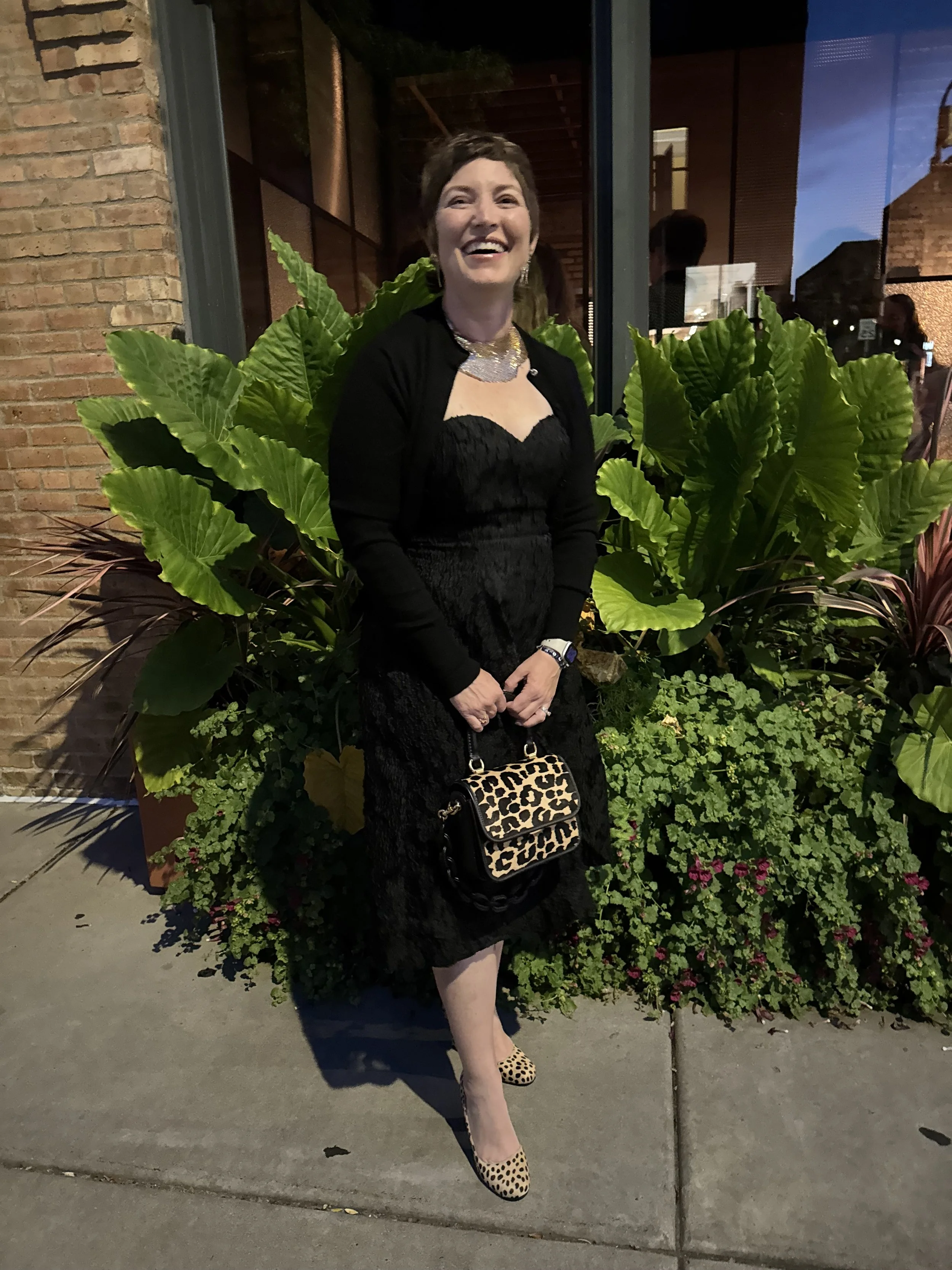 A woman smiling and standing outside at night, wearing a black dress with a black cardigan, leopard print shoes, and holding a leopard print purse.