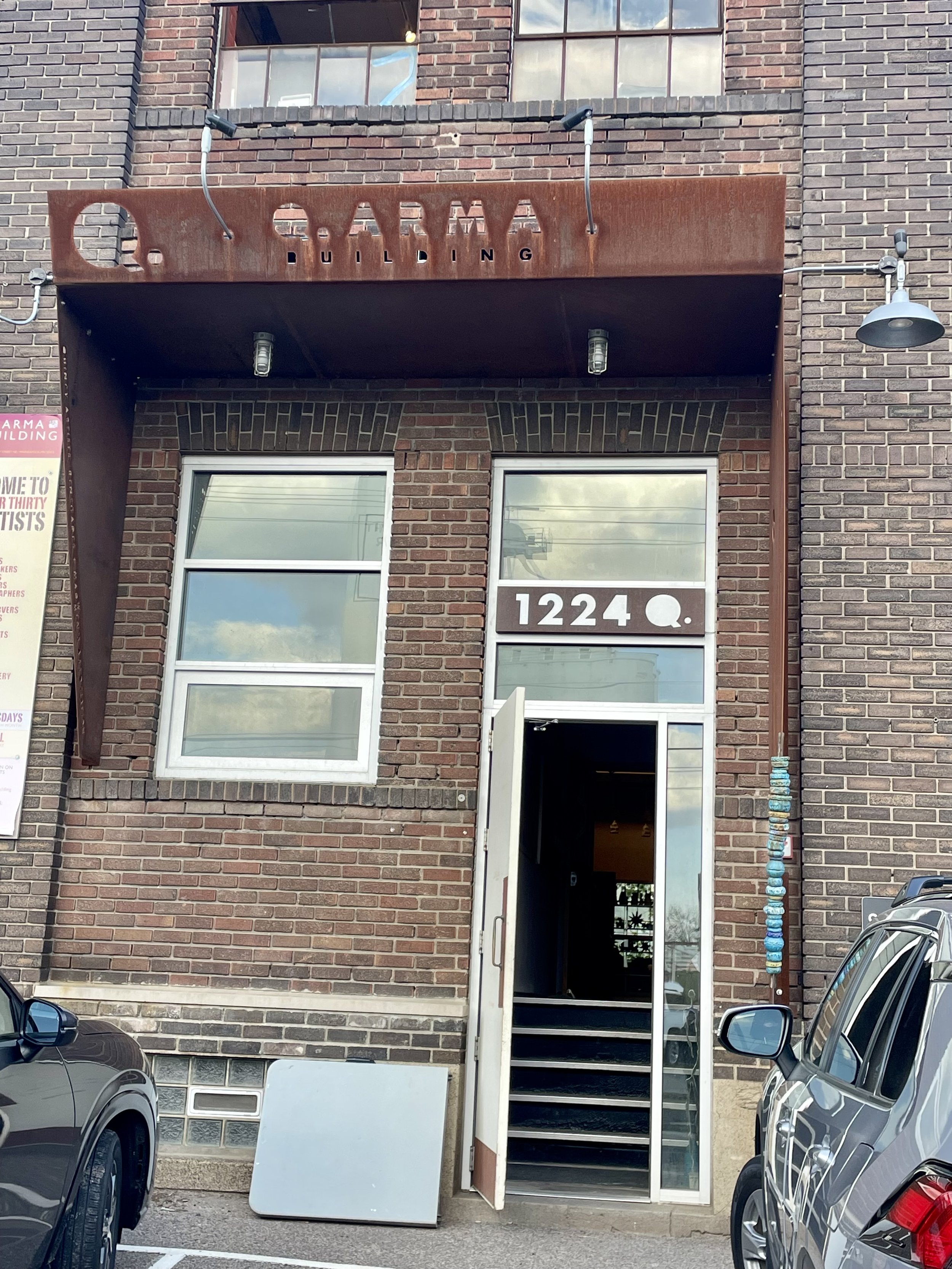 Exterior of a brick building with a sign reading 'QARMA BUILDING' and the address '1224 Q.' The entrance has a door with stairs leading inside, and a string of decorated blue and white beads hanging beside it. Cars are parked in front.