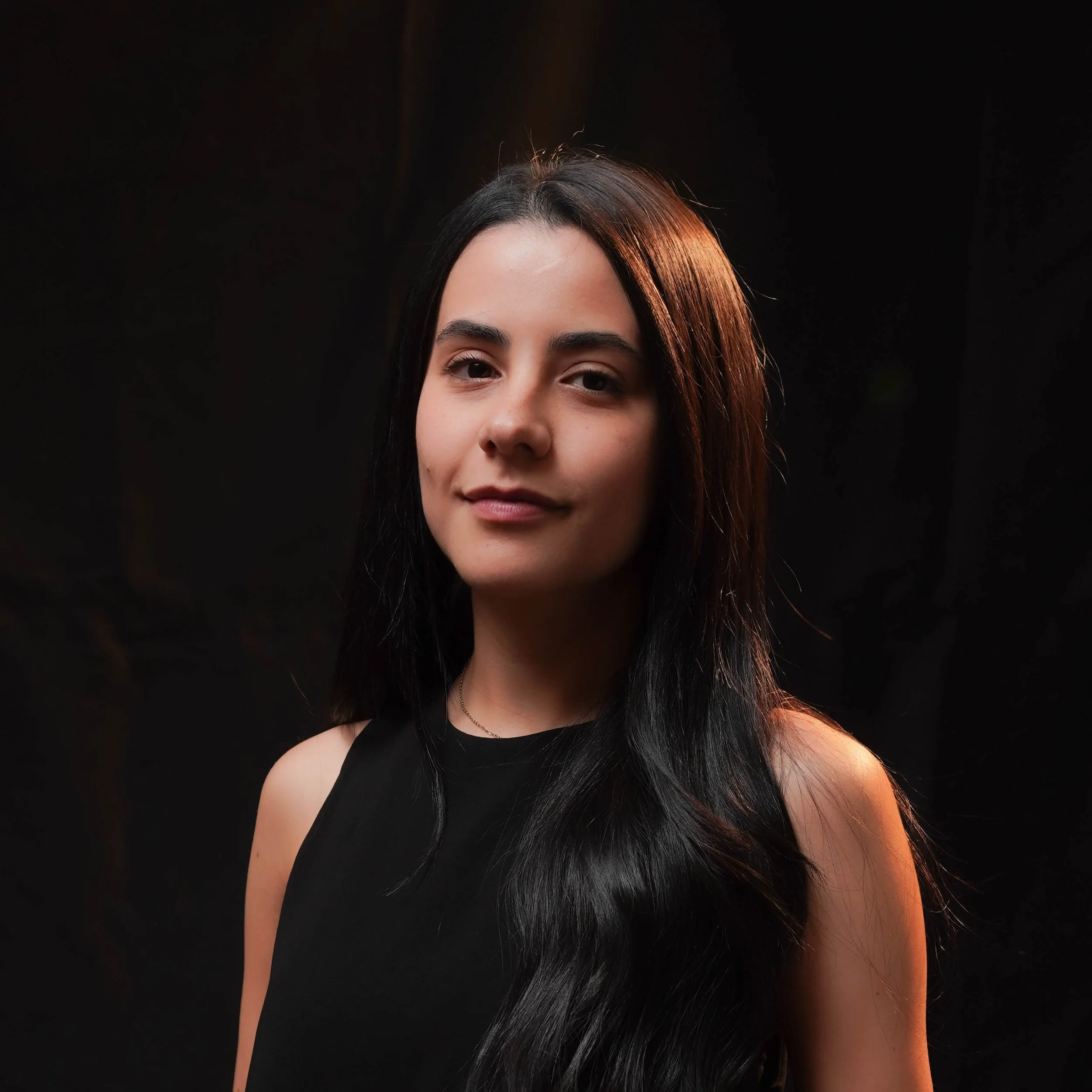 Close-up portrait of a young woman with long dark hair, wearing a black sleeveless top, against a dark background with warm lighting highlighting her profile.