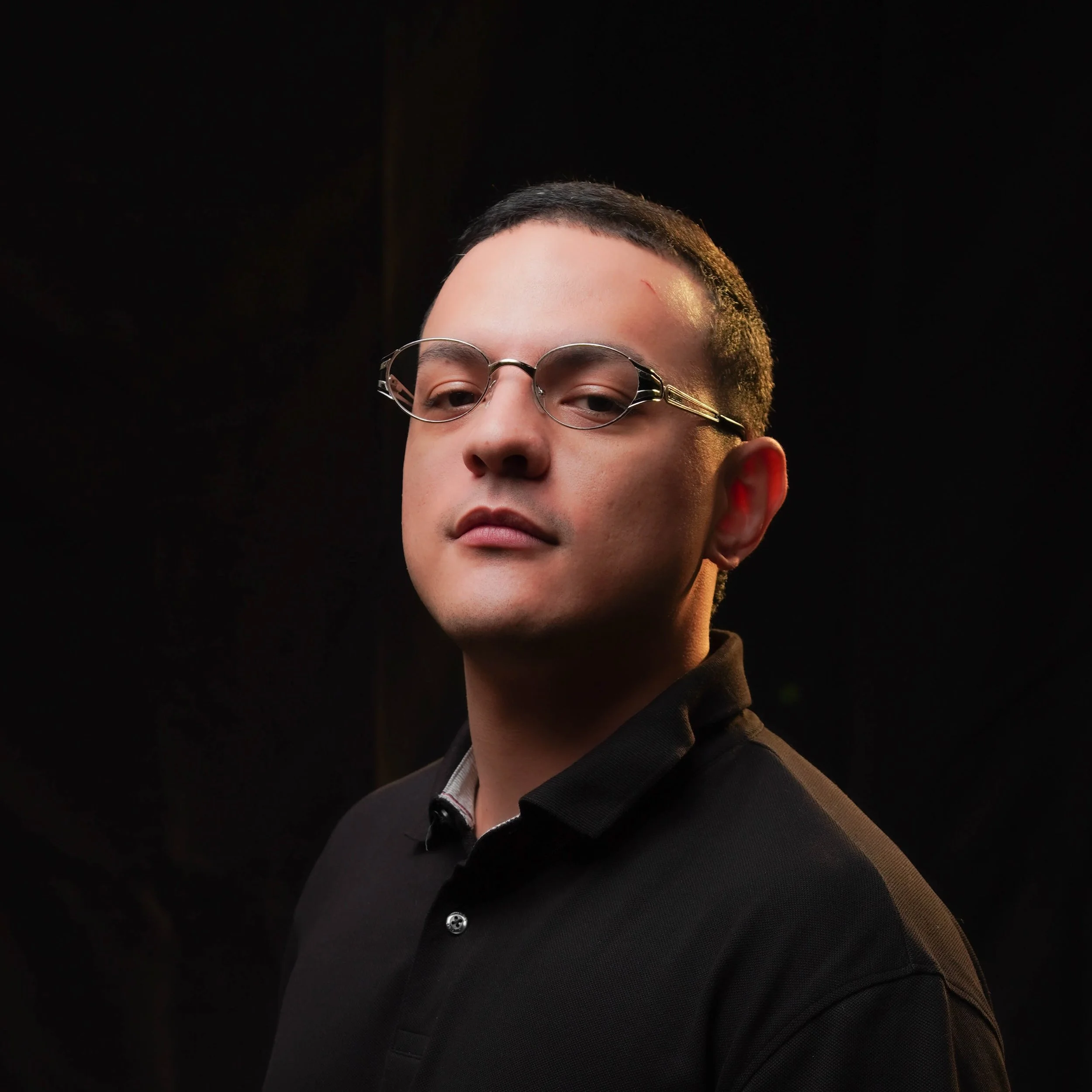 Close-up of a person with short black hair and round glasses, wearing a dark blazer and shirt, against a black background.