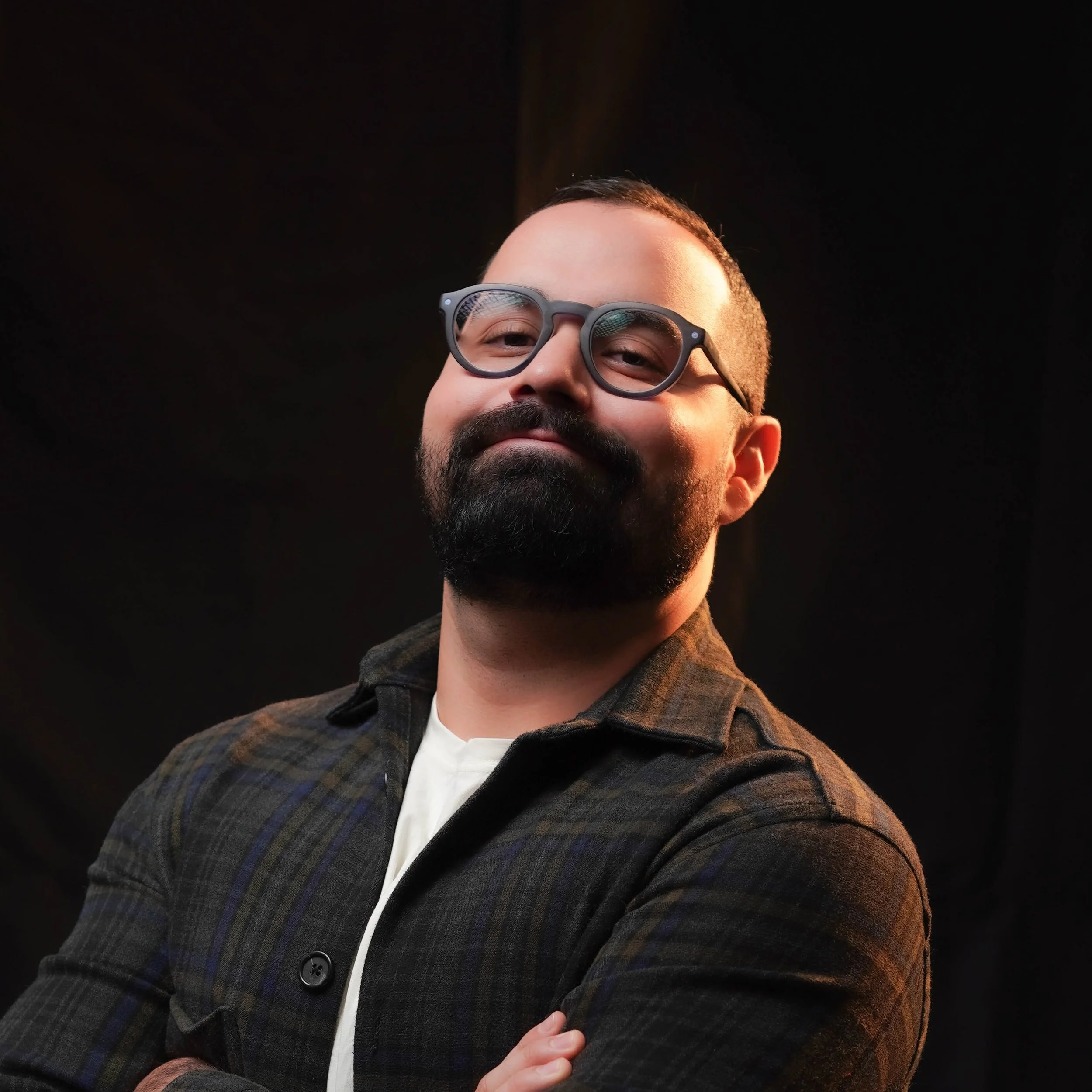 A man with a beard and glasses, wearing a plaid jacket, smiling while holding his chest, against a dark background.