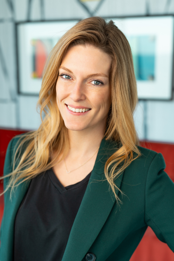 A woman with long blonde hair smiling at the camera, wearing a dark top and green blazer, in an office environment with monitors in the background.
