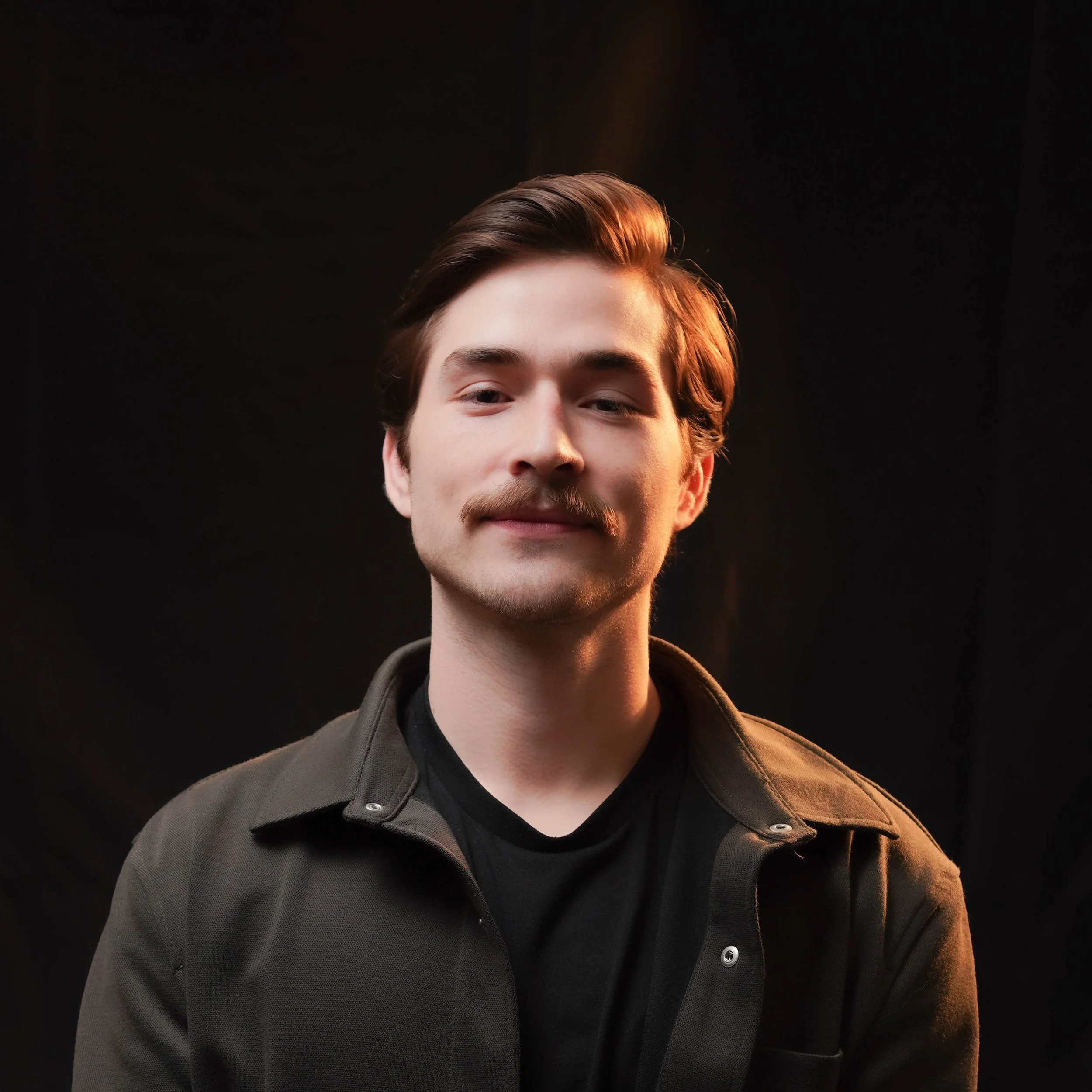 Close-up of a young man with brown hair, a mustache, and beard, wearing a dark jacket against a black background.