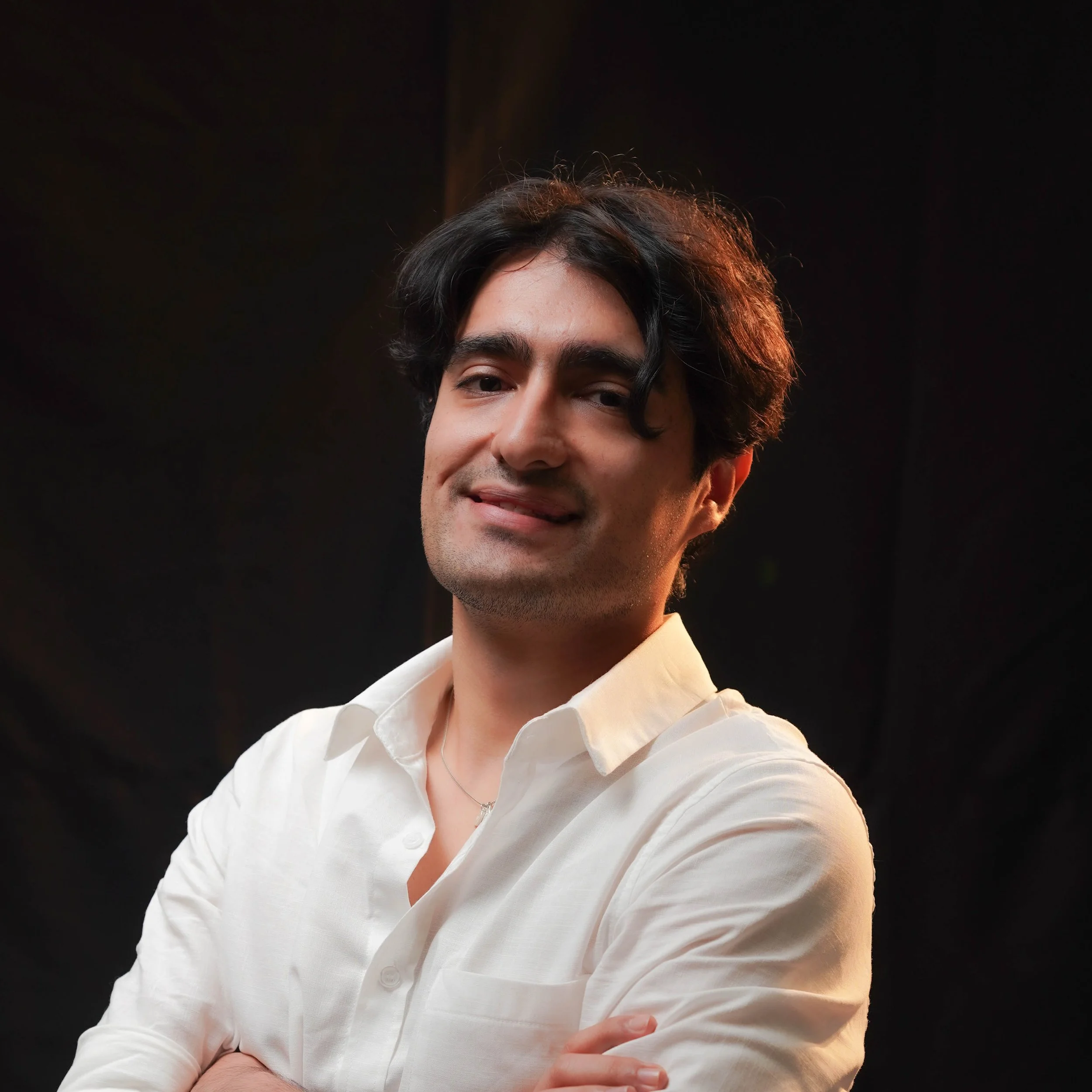 Close-up of a man with dark, wavy hair wearing a white shirt, smiling with arms crossed, against a black background.