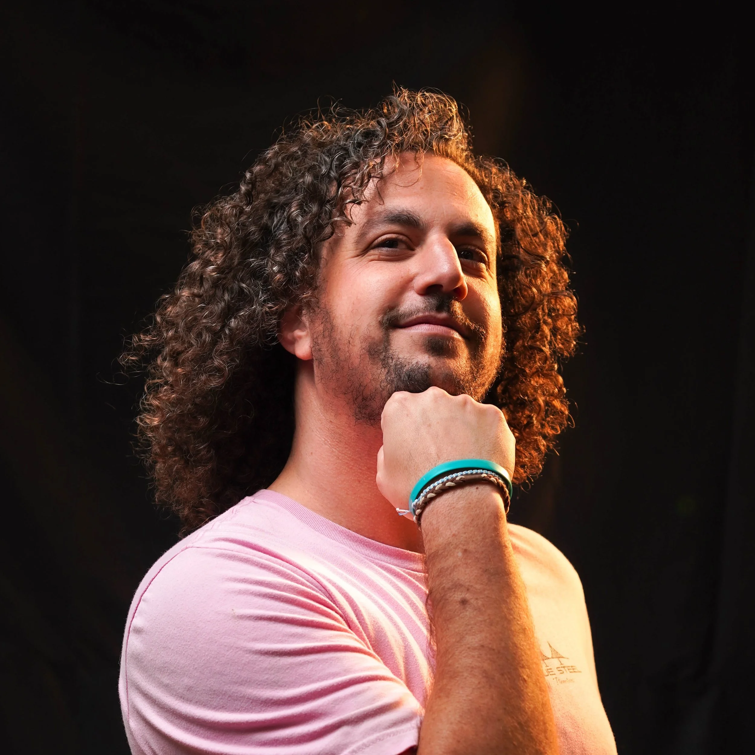 A person with curly hair wearing a pink shirt and a teal bracelet, smiling and resting their chin on their hand, against a dark background.