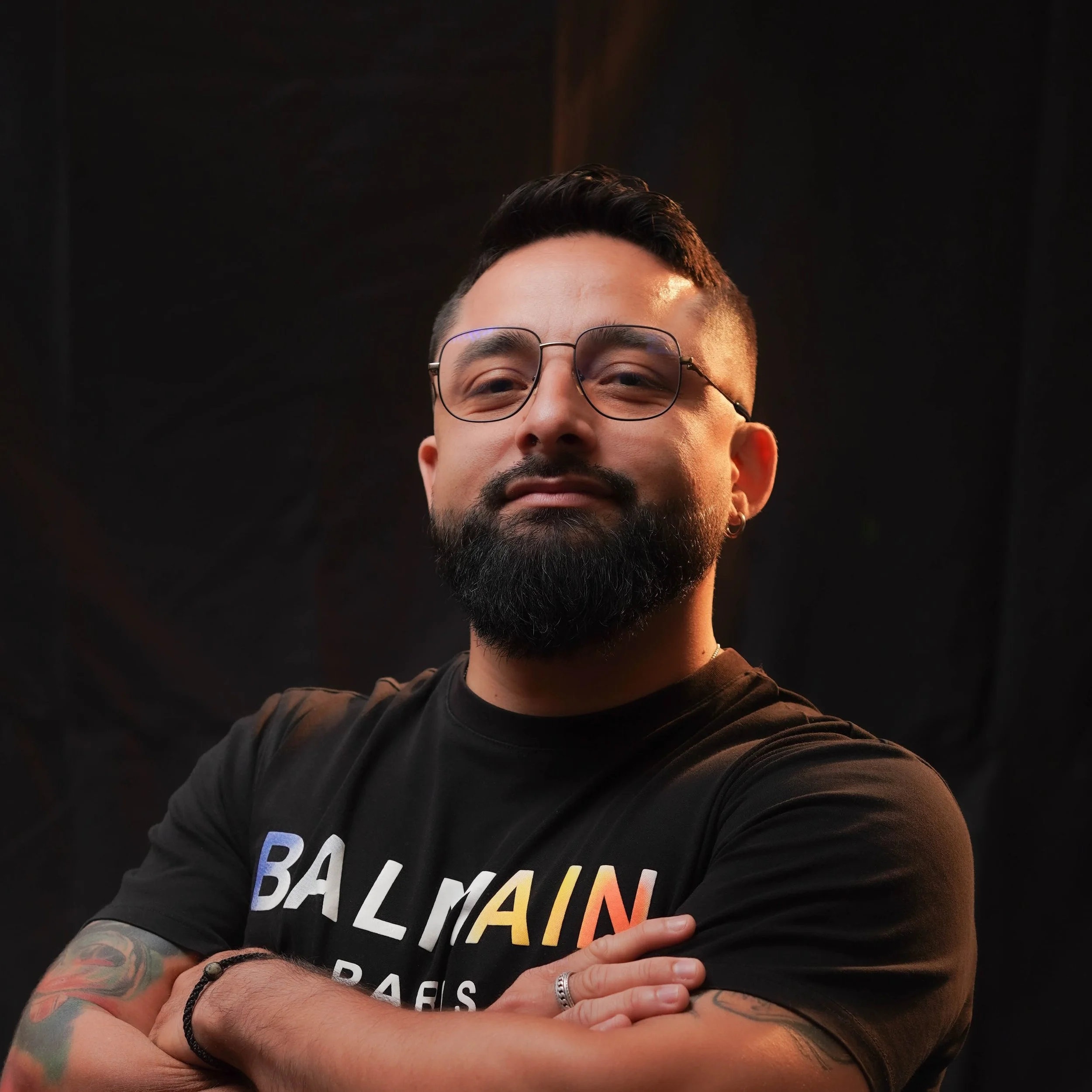 Part of a man's face with glasses and tattoos on arm, wearing a black t-shirt with colorful text, standing against a dark background.