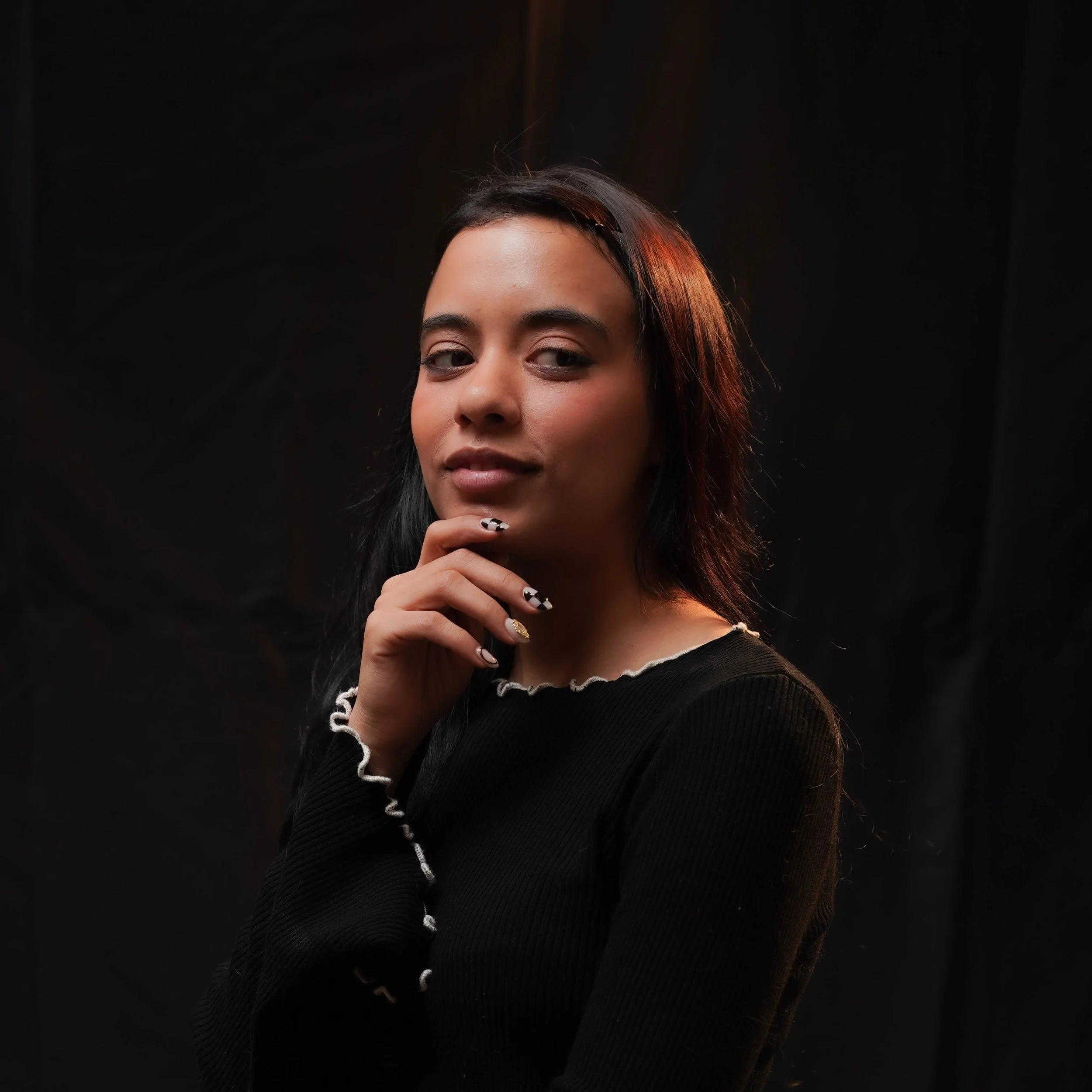 A woman with dark hair and makeup, wearing a black top with white trim, is touching her chin with her hand, showing her rings and nail art, against a dark background.