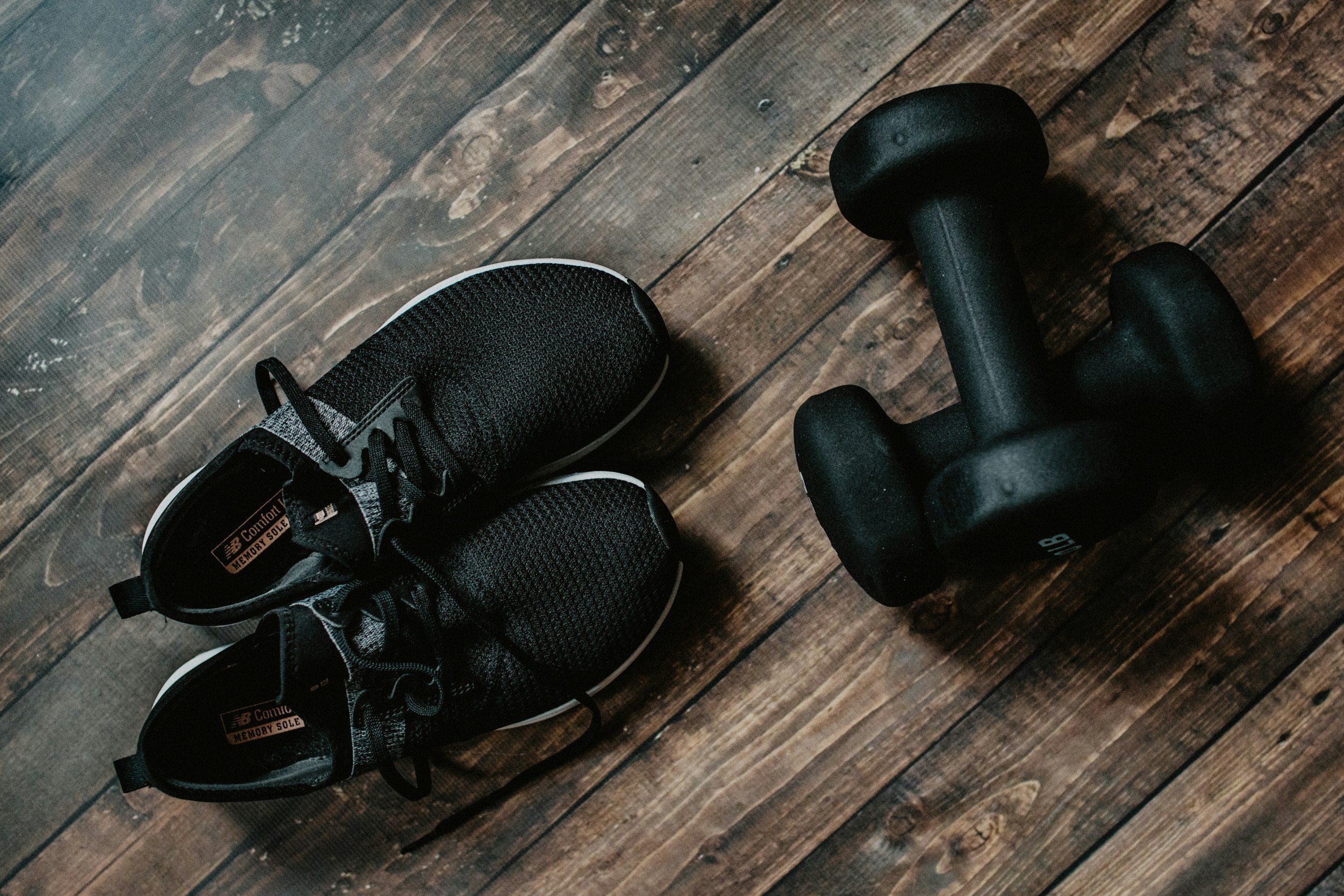 Pair of black athletic shoes and a black dumbbell on a wooden floor.