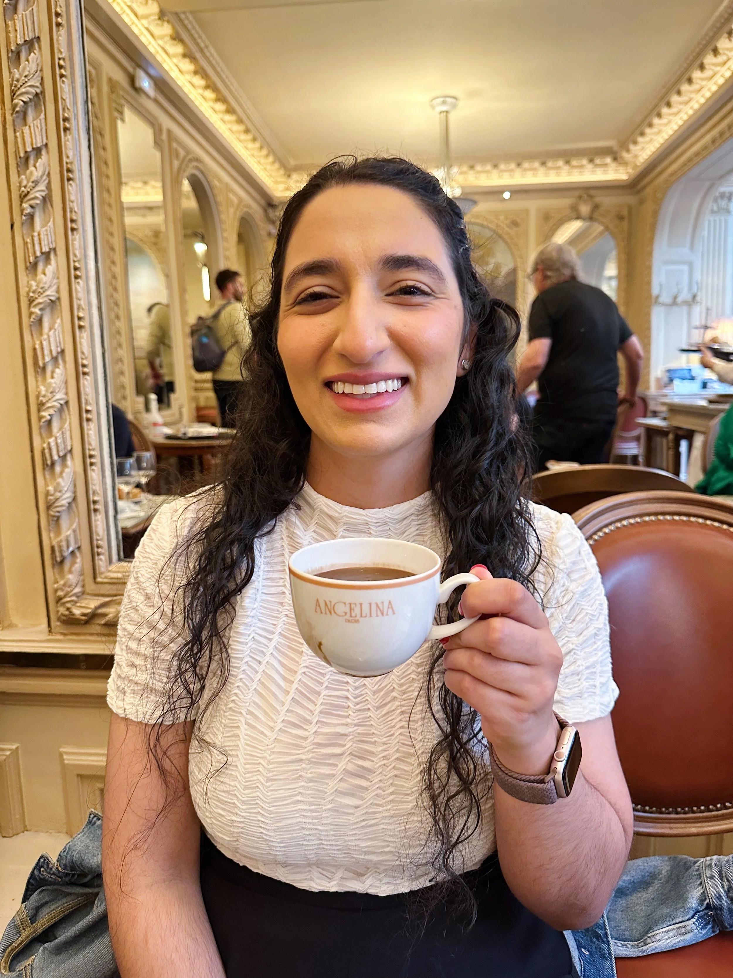A woman with long dark curly hair smiling and holding a cup of coffee in a fancy, ornately decorated cafe. She is wearing a white textured top and a smartwatch. The cafe has gold accents, large mirrors, and other patrons in the background.