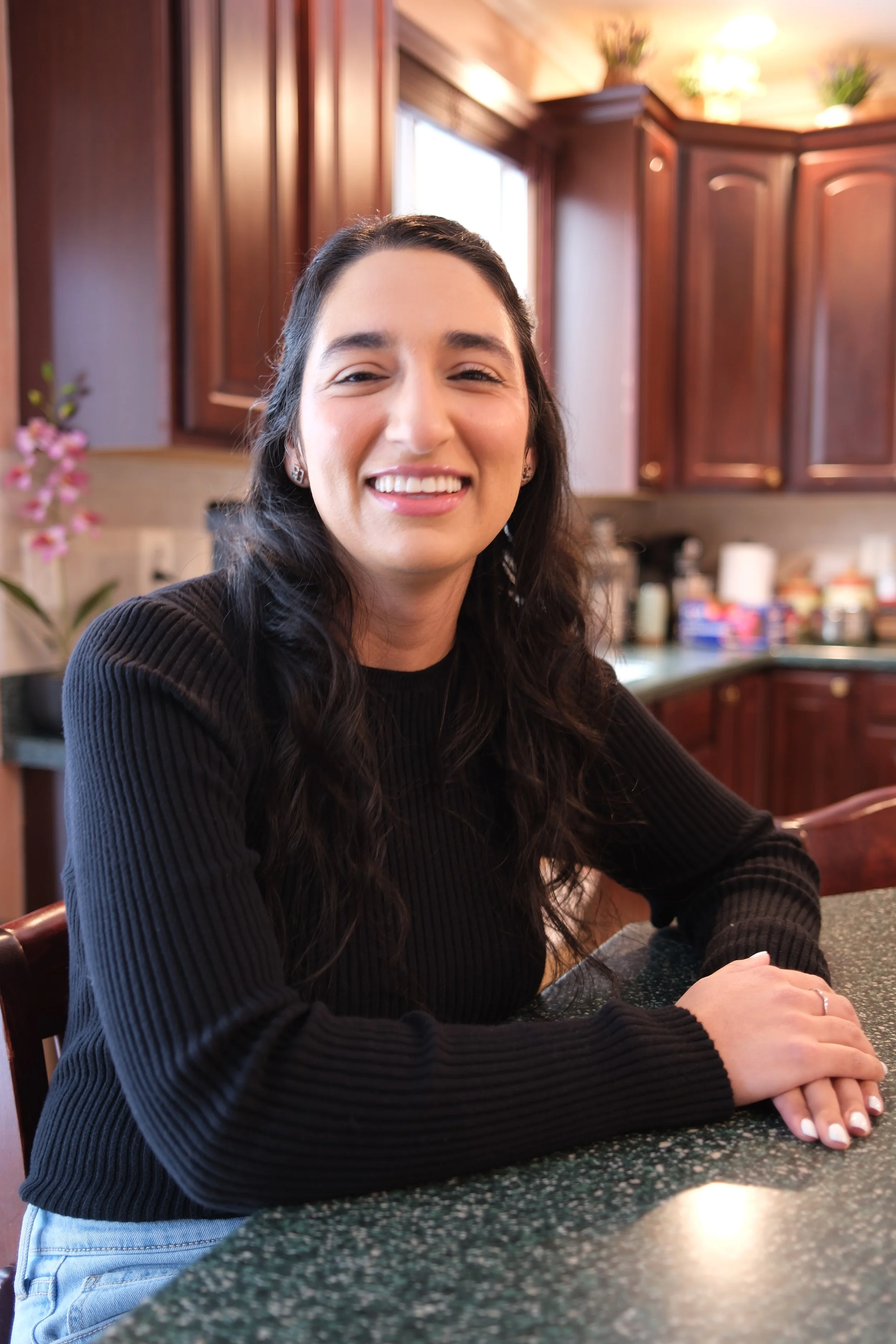 A woman with long dark hair and a black sweater sitting at a kitchen table, smiling at the camera.