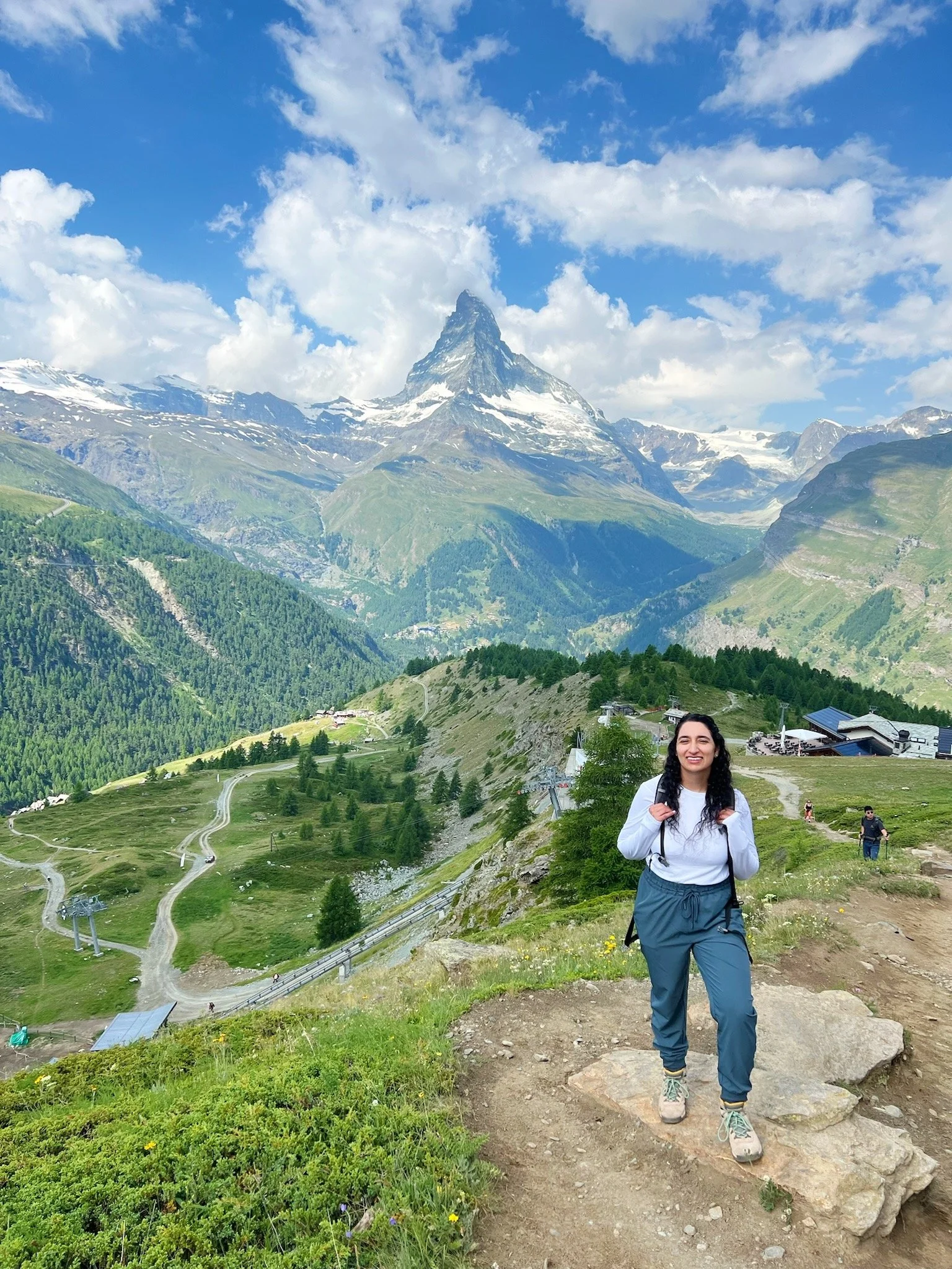 A woman hiking in the mountains with a trail and green landscape behind her, with snowy peaks and a partly cloudy sky