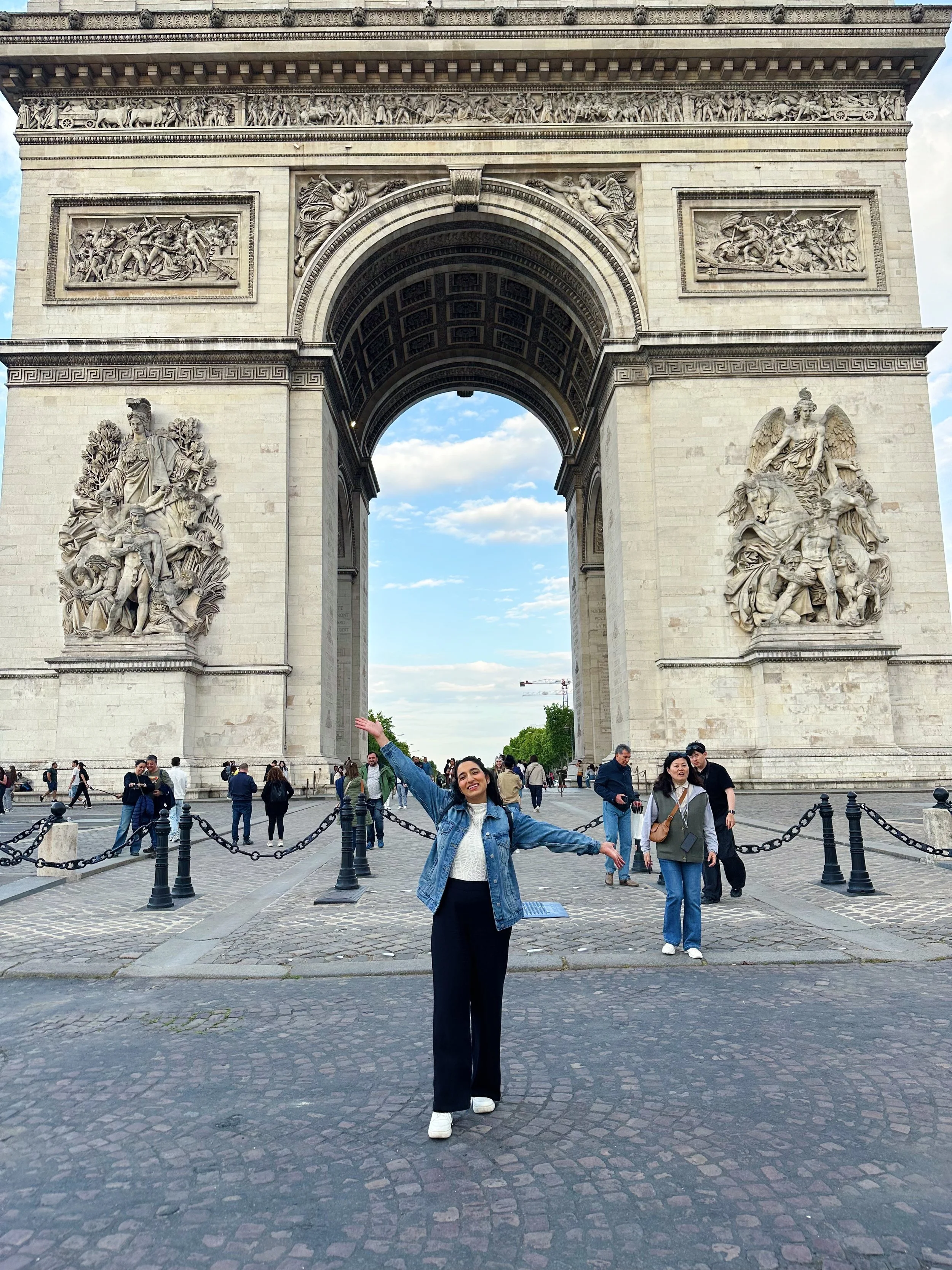 A woman standing with arms outstretched in front of the Arc de Triomphe in Paris, France, with other tourists around and a partly cloudy sky above.
