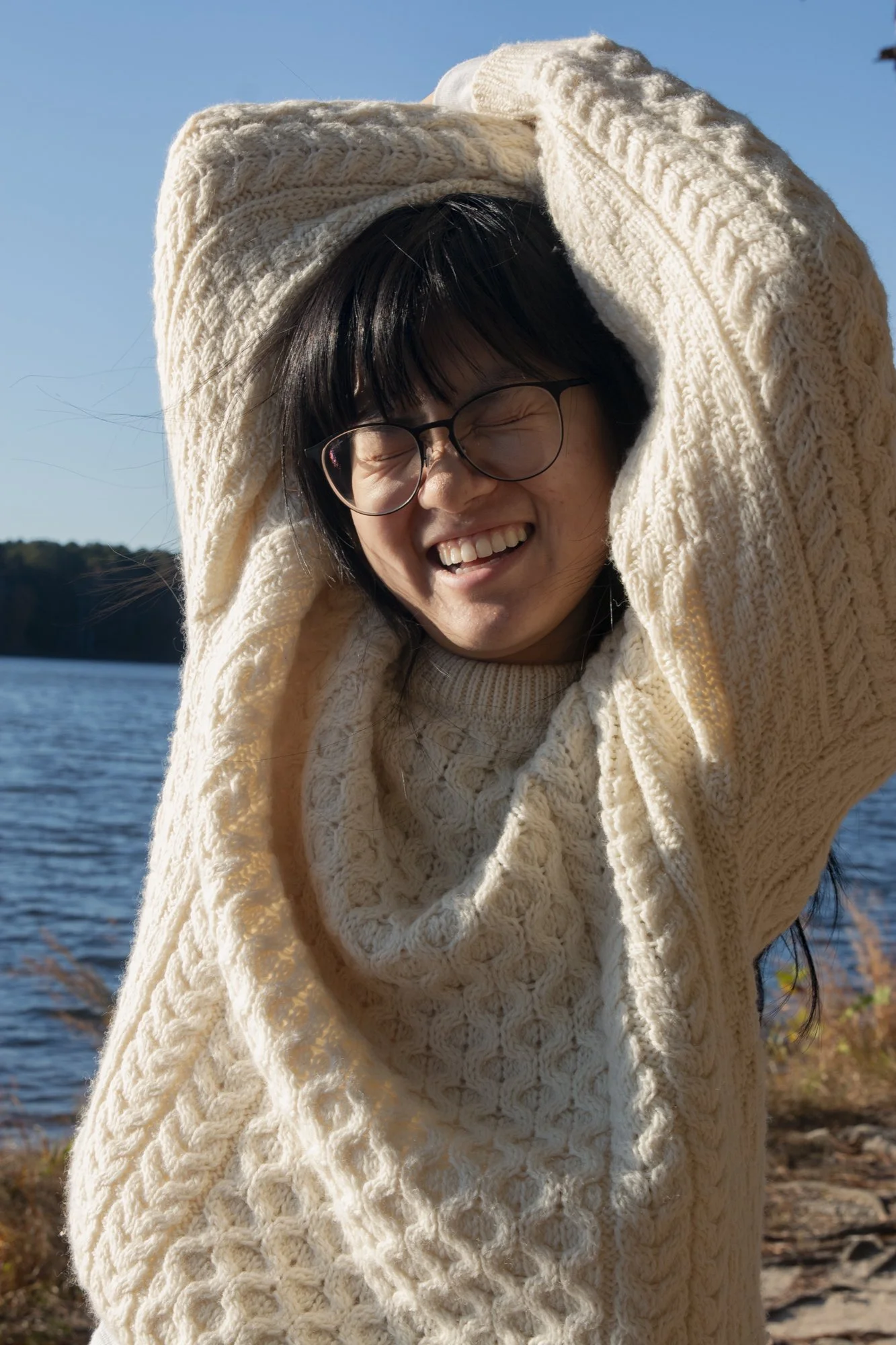 A woman with black hair, glasses, and a cream cable-knit sweater smiling with her eyes closed by a lake on a sunny day.