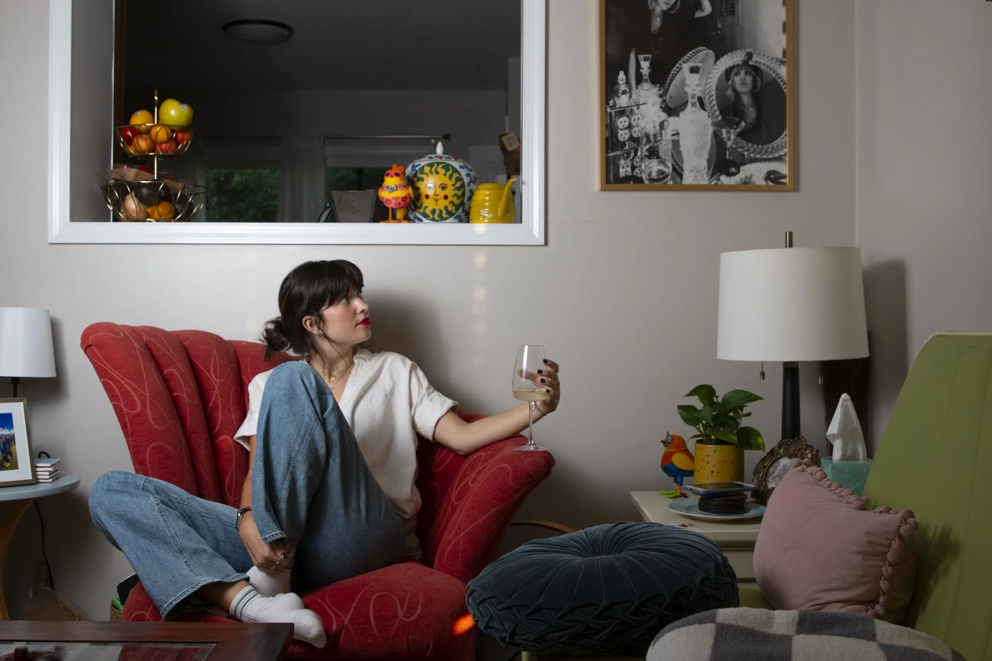 A woman with dark hair tied back, sitting on a red armchair in a living room, holding a glass of wine, wearing a white shirt, blue jeans, and white socks. The room has beige walls, a white side table with framed photo, a lamp, and colorful decoration