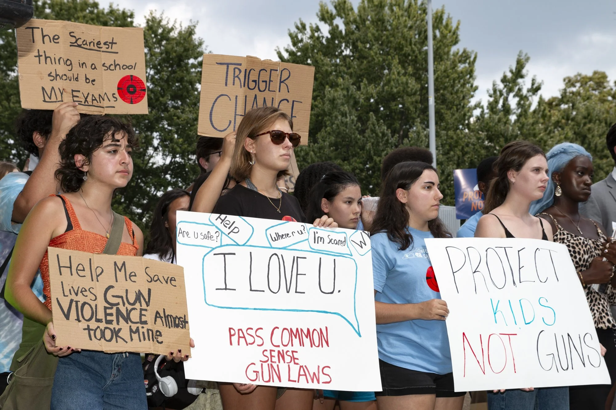Group of young women protesting, holding signs advocating for gun control and safety, with trees and cloudy sky in background.
