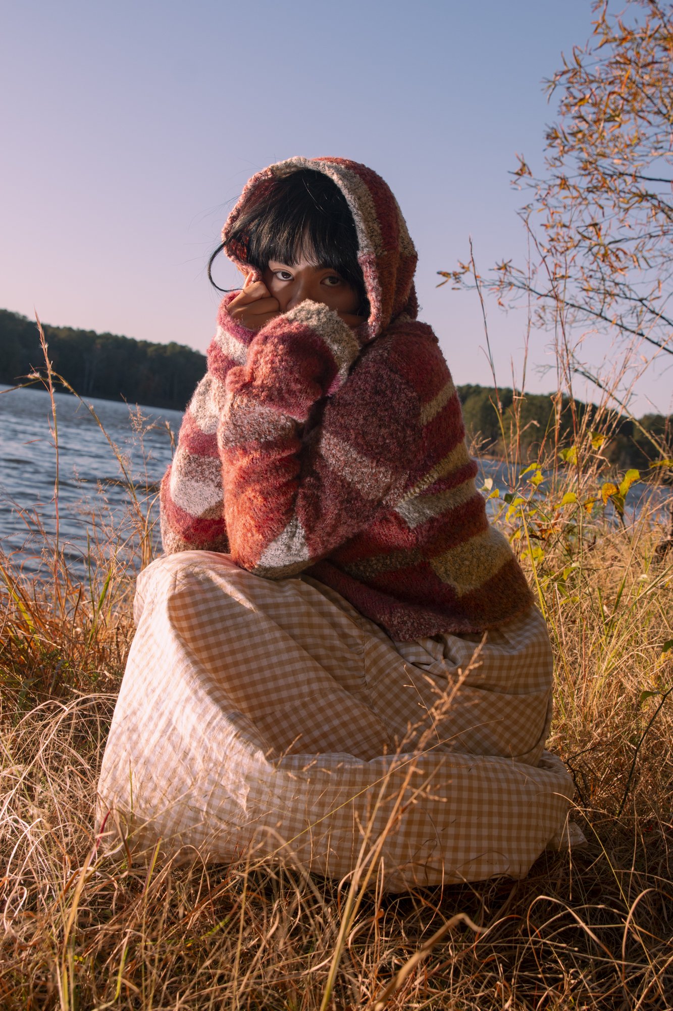 A person sitting on grass by Jordan Lake during sunset, wearing a hooded, red and beige patterned sweater and beige checkered skirt, with one hand near their face and partially covering their mouth.