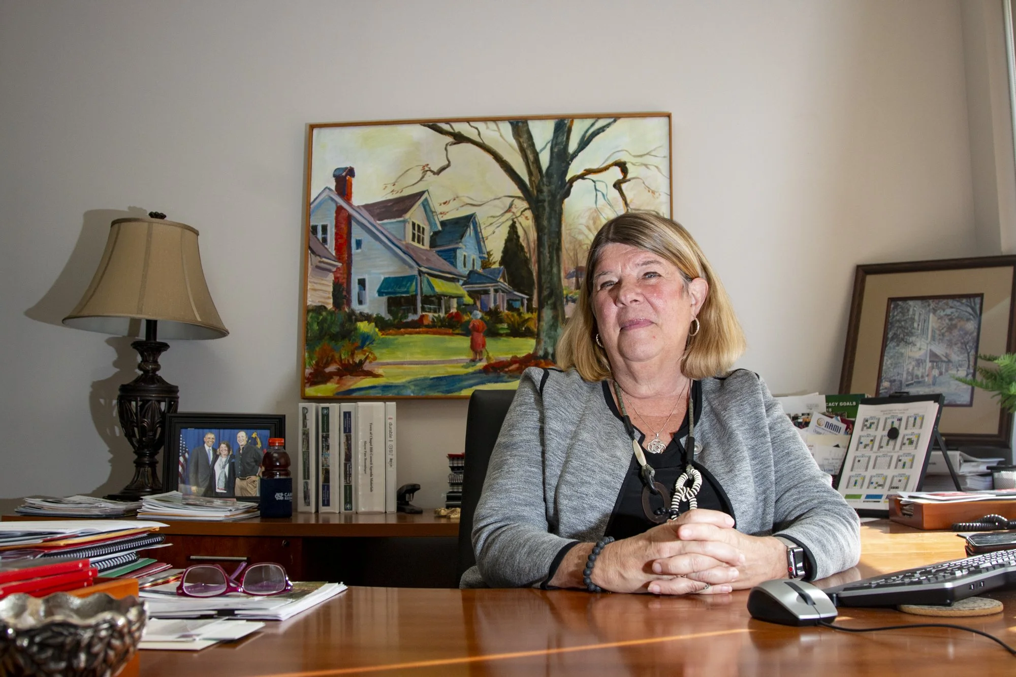 Mayor Pam of Chapel Hill, NC sitting at a cluttered wooden desk in her office, with framed photos, books, and a lamp in the background, and paintings on the wall.
