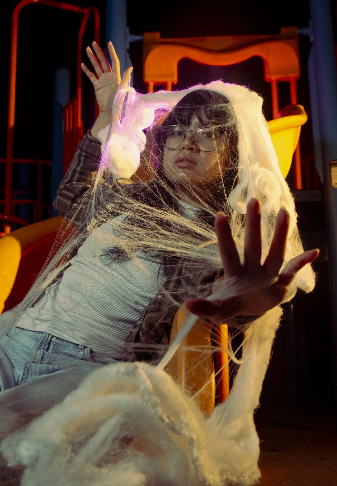 A person with glasses and dark hair is sitting on a slide covered with fake spider webs. The person raises one hand near their face and the other hand extended forward as if reaching out. The background features playground equipment illuminated with 