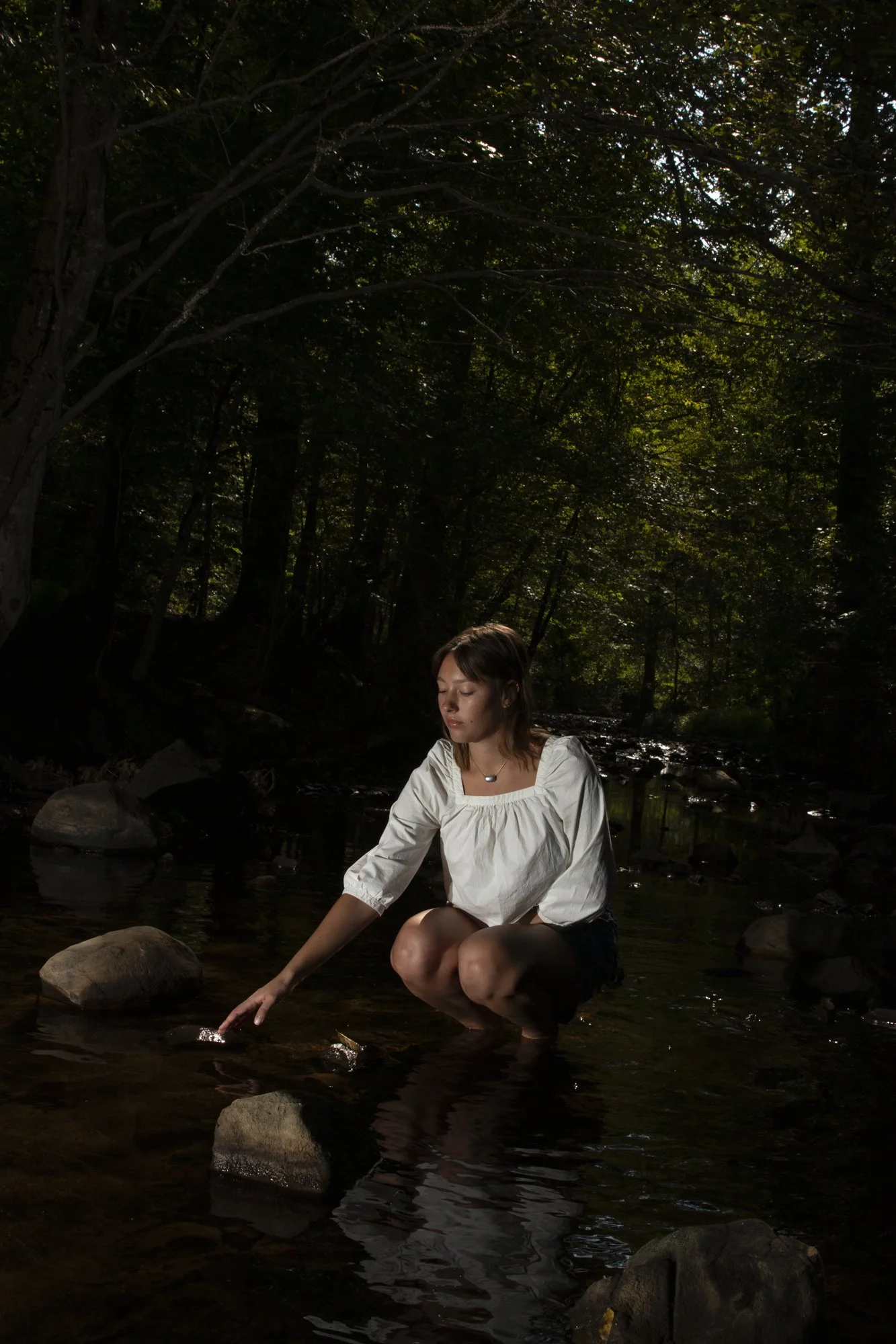 A young woman kneeling in a shallow creek in a wooded area, touching the water with her hand, with sunlight filtering through trees.