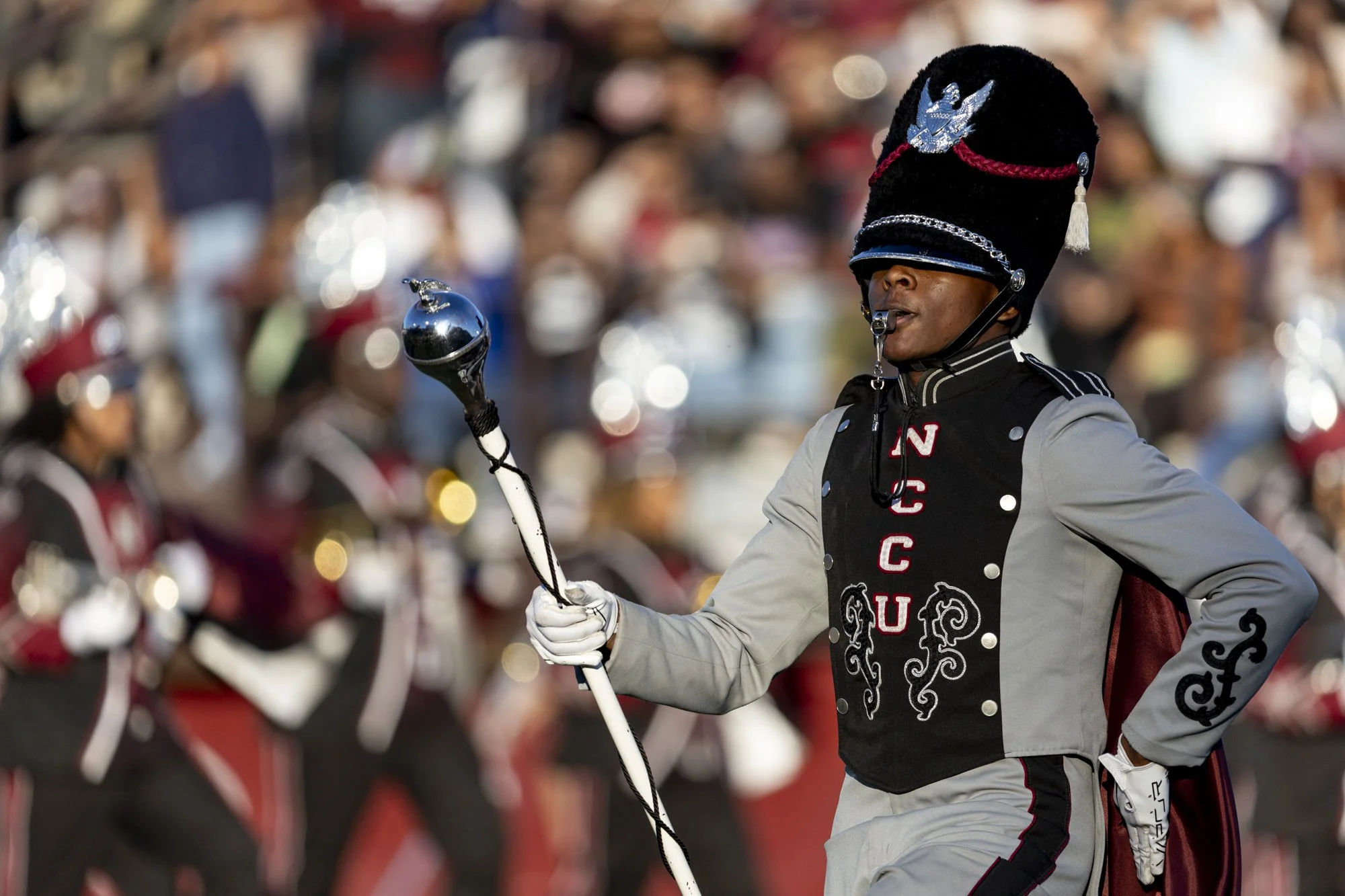 A marching band member for NC Central dressed in a black and red uniform and large black hat with a tall plume, holding a baton, during a parade or performance with a blurred crowd in the background.
