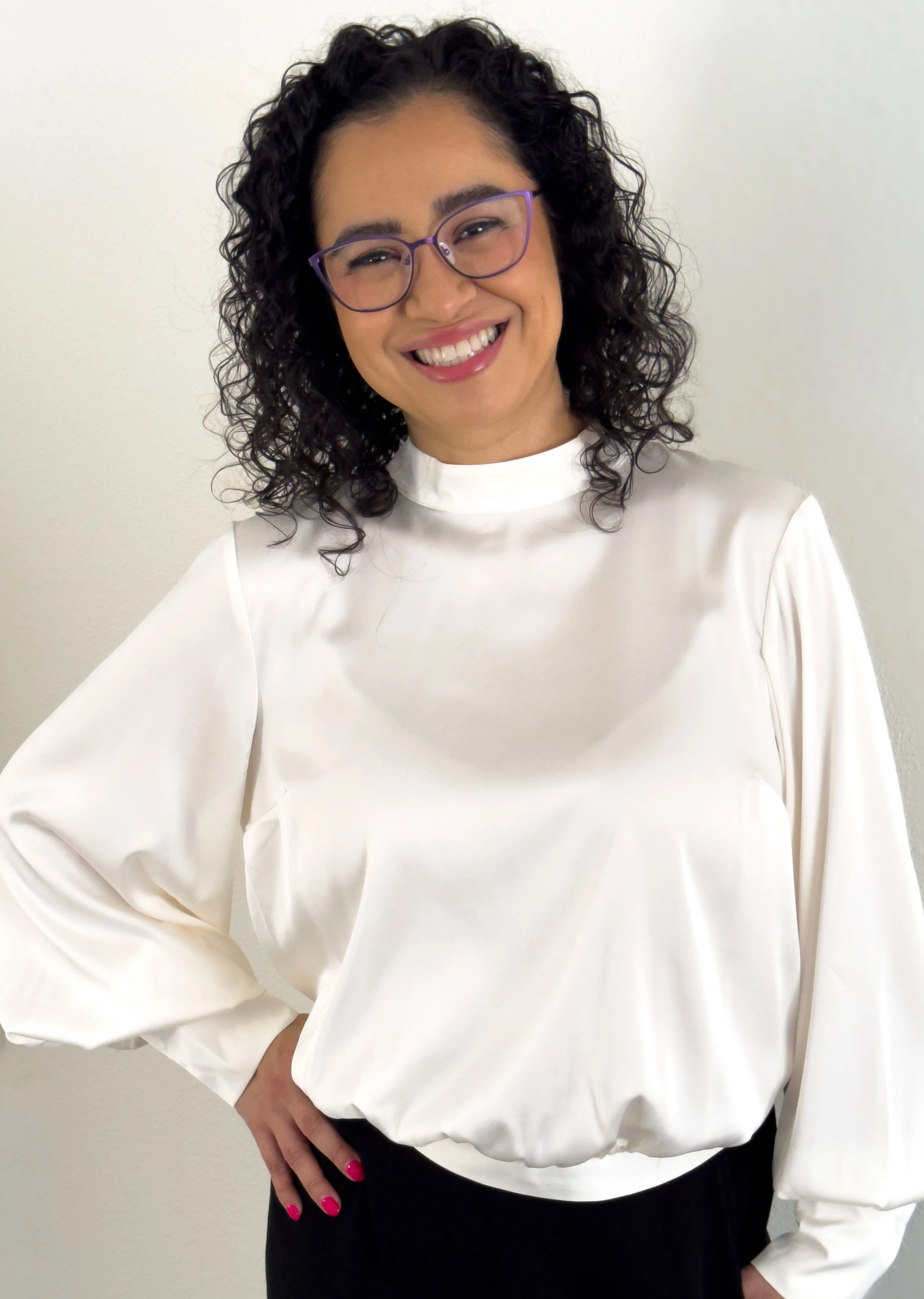 Mujer sonriente con gafas moradas, cabello rizado, vestida con blusa blanca y pantalón negro, posando con una mano en la cintura frente a una pared blanca.