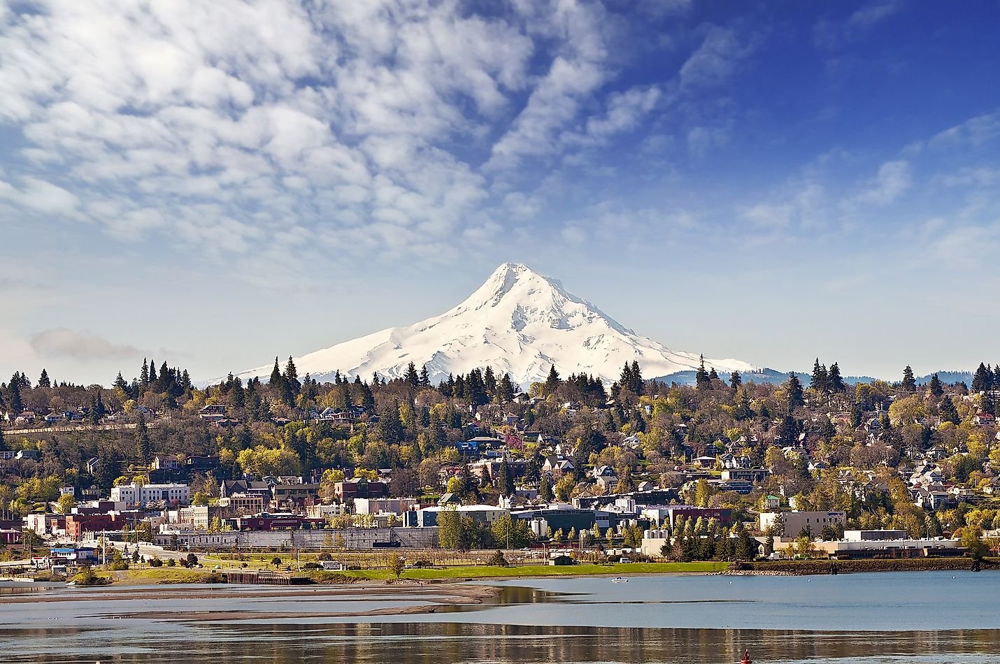 Vista del volcán Mount Hood cubierto de nieve con una ciudad y un río en primer plano, bajo un cielo parcialmente nublado.