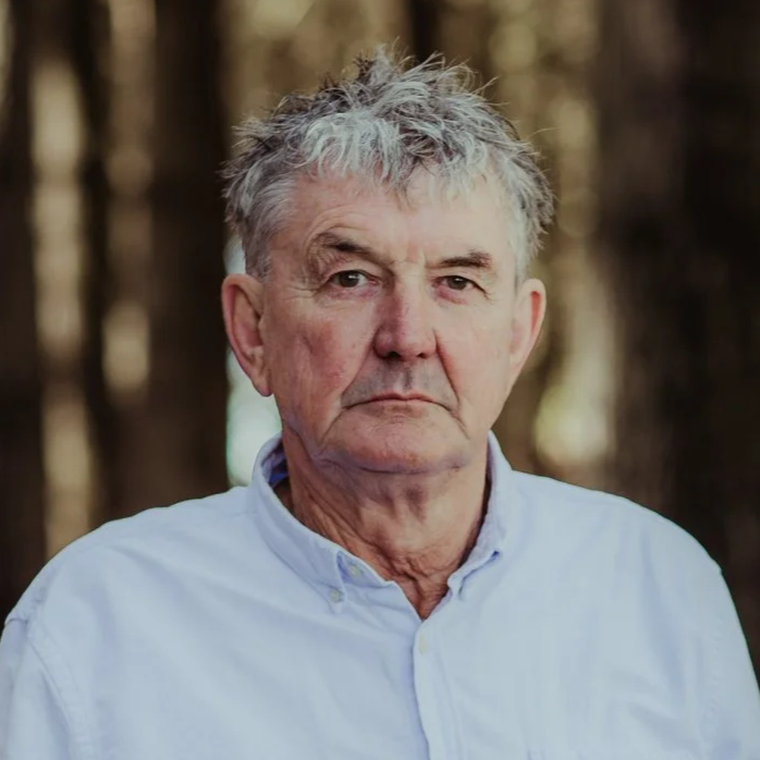A serious-looking elderly man with gray hair, wearing a white button-up shirt, standing outdoors with a blurred background of trees.