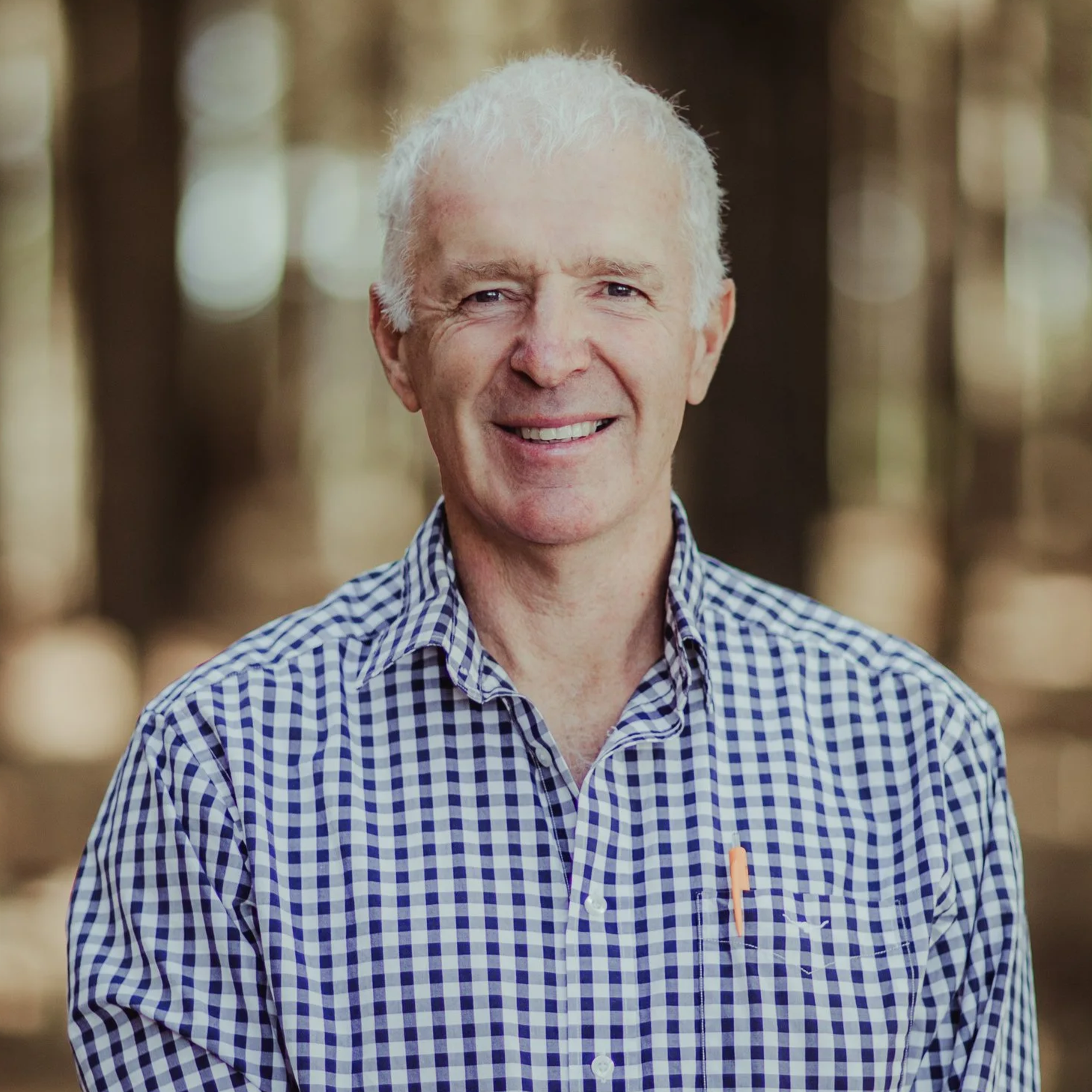 A smiling older man with white hair wearing a blue checkered shirt, standing outdoors in a wooded area.