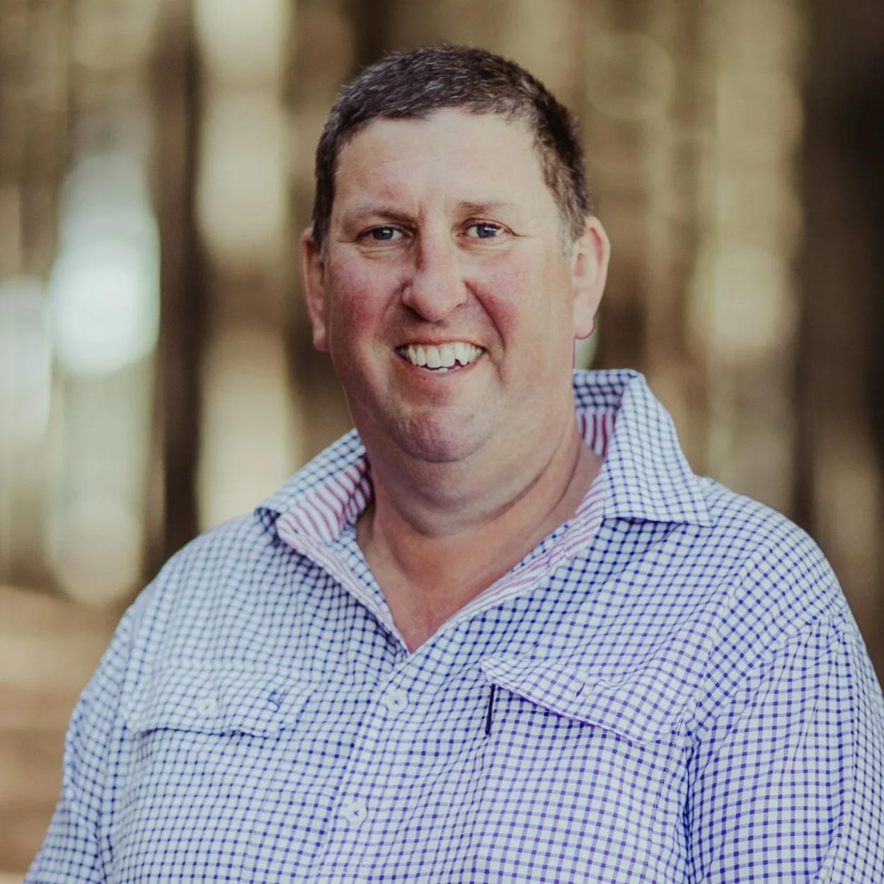 A smiling man with short dark hair wearing a checkered button-up shirt outdoors with a blurred background of trees.