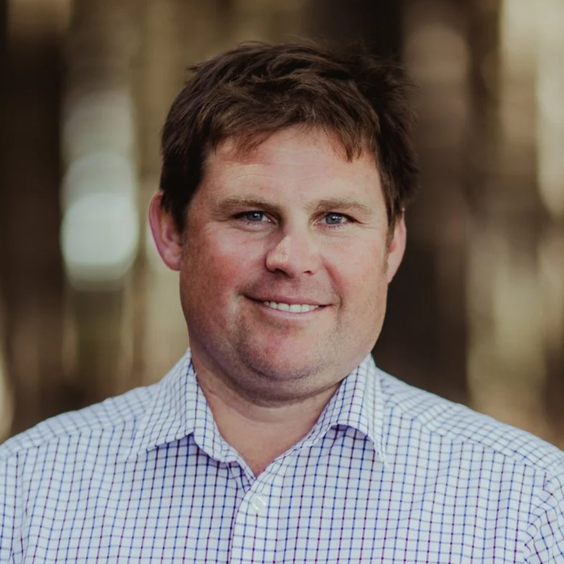 A man with brown hair and blue eyes wearing a white checkered shirt, smiling outdoors with blurred trees in the background.