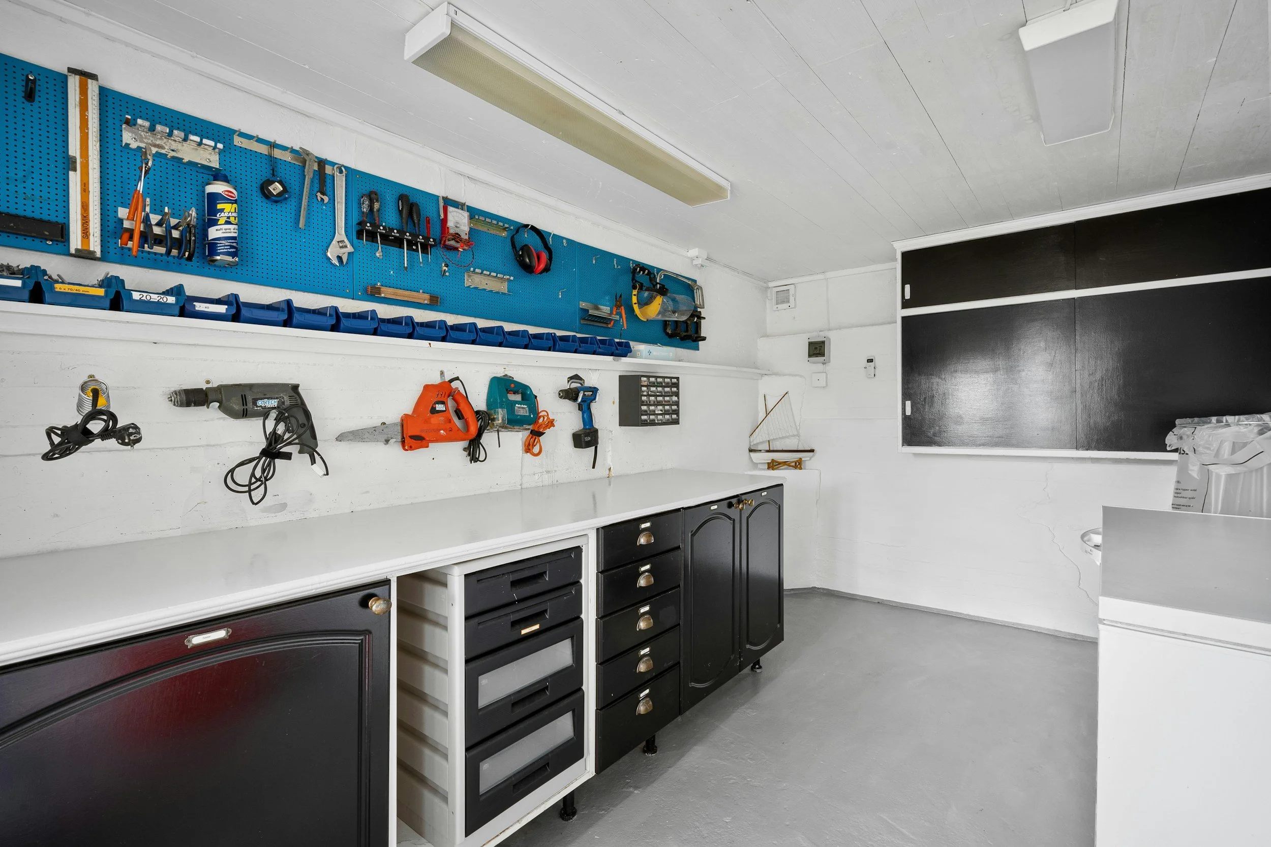 A well-organized workshop with tools hanging on a blue pegboard, power tools on a white countertop, and cabinets underneath. There is a large black chalkboard on the wall and a gray concrete floor.