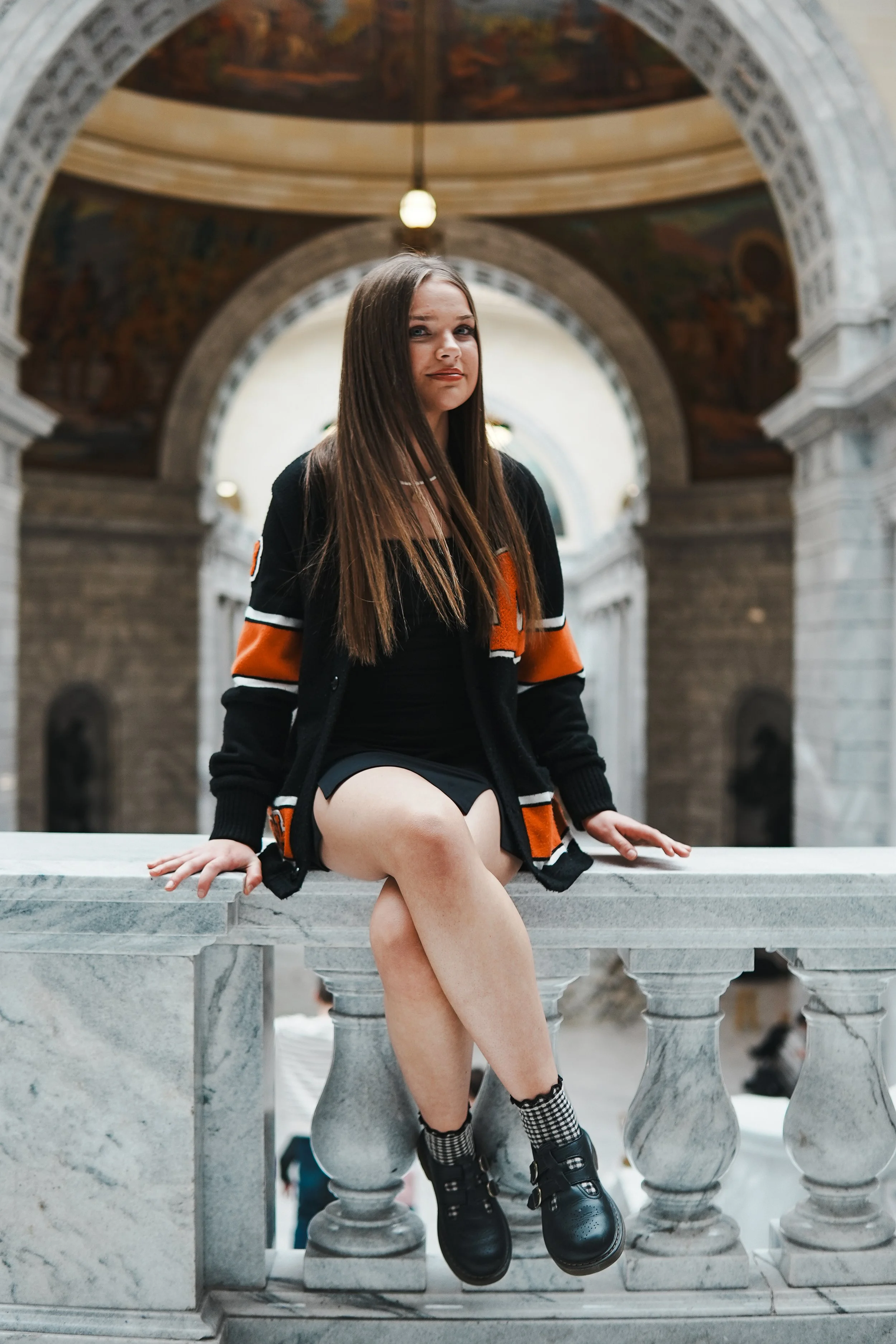A young woman with long brown hair sitting on a marble railing inside a historic building with arches and murals.
