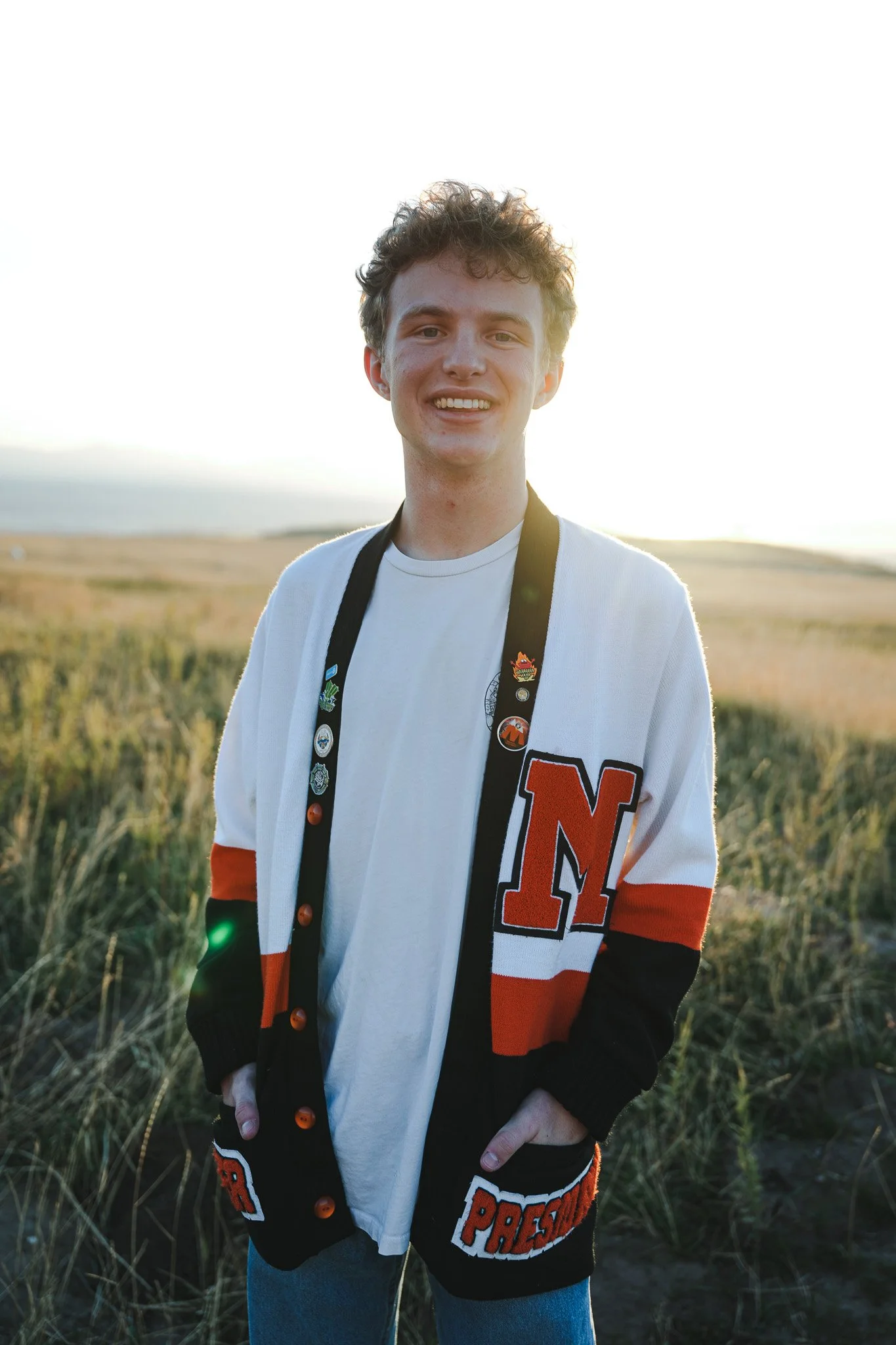 A young man standing outdoors in a grassy field during sunset, smiling, wearing a varsity-style jacket with an 'N' on it and pins on his lanyard.