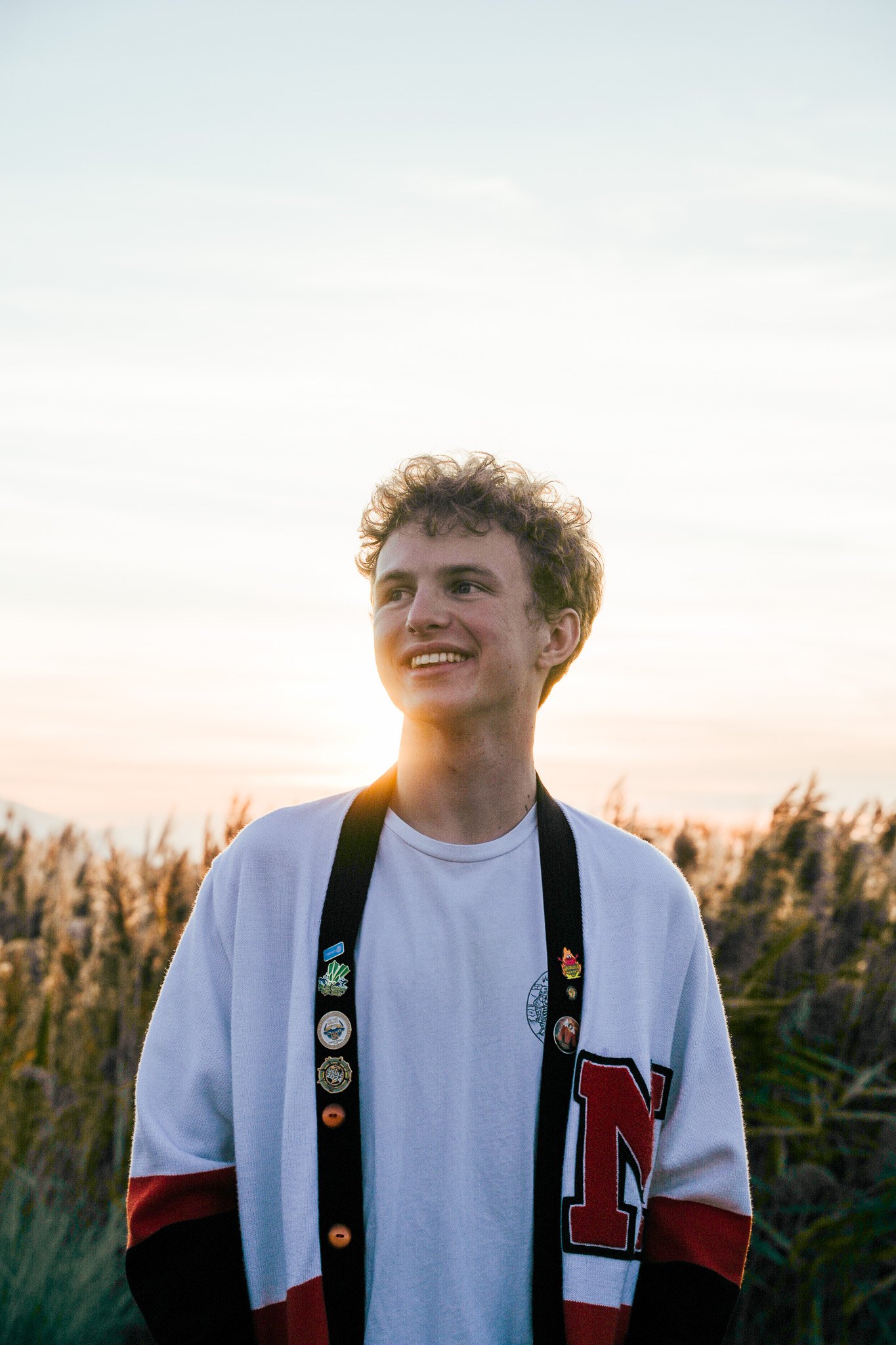 A young man with curly hair smiling outdoors during sunset, wearing a white sports jersey with red and black lettering and a lanyard with pins around his neck.