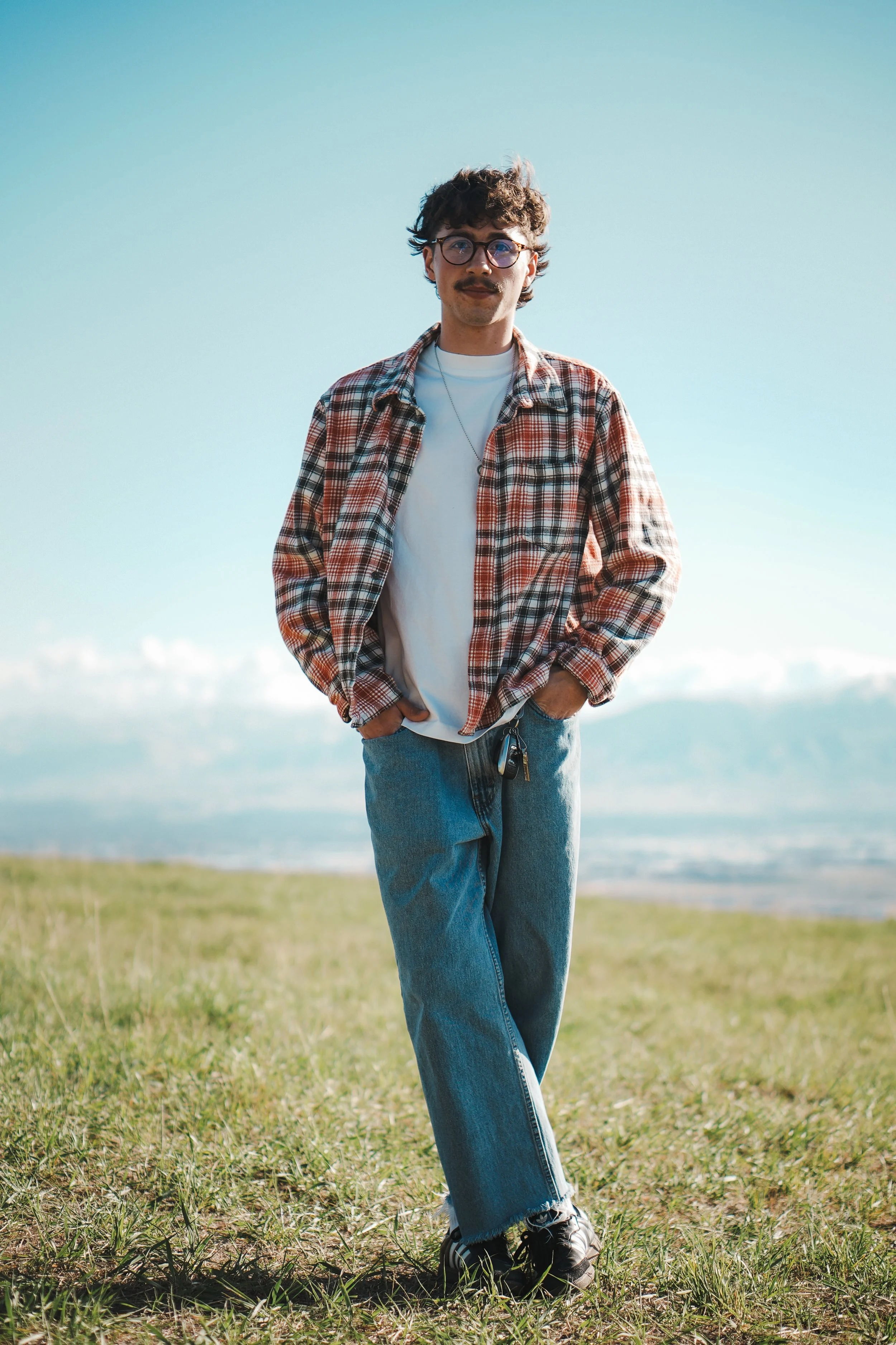 A young man with curly hair and glasses standing outdoors on grass near the ocean, dressed in a plaid shirt, white t-shirt, blue jeans, and sneakers.