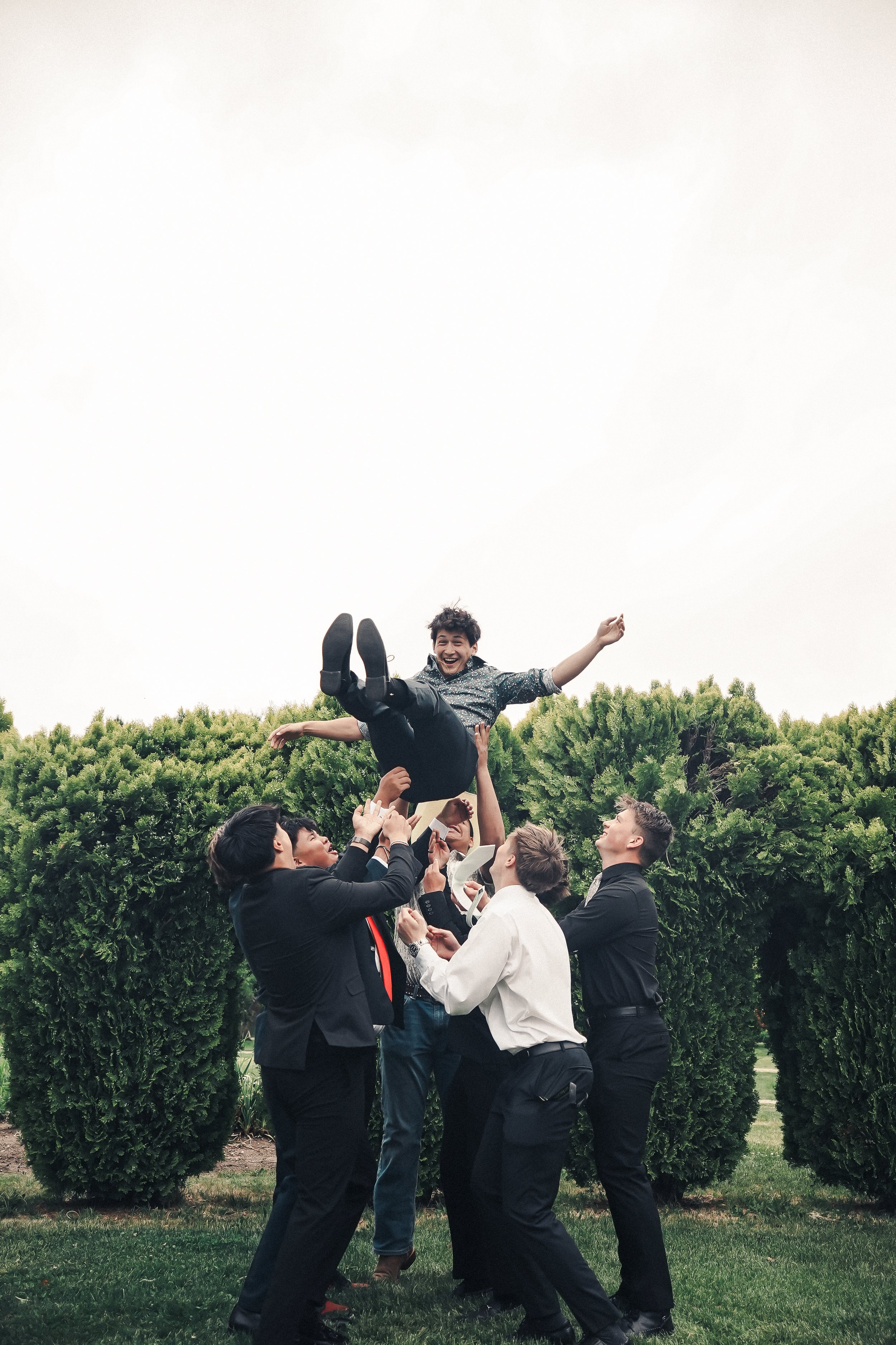 A group of people in business attire joyfully lifts a young man into the air outdoors with greenery and bushes in the background.