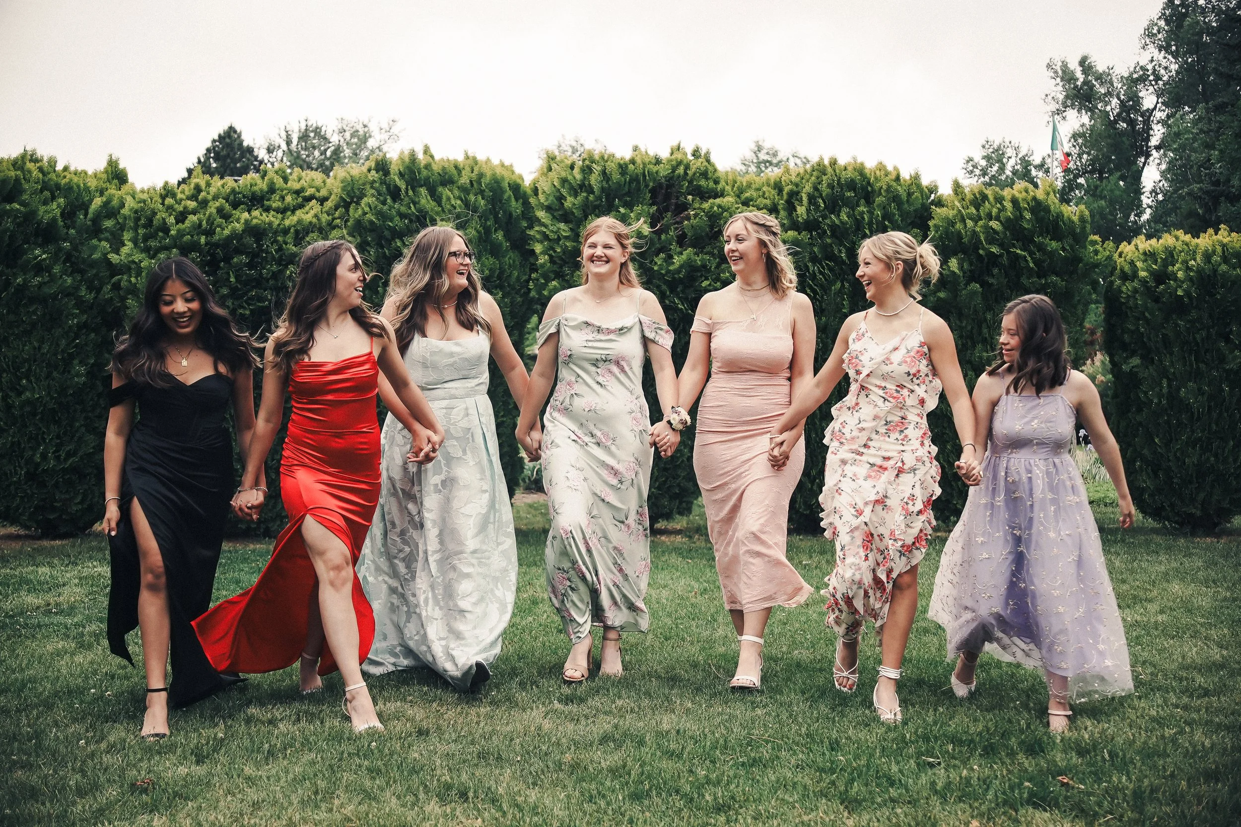 A group of eight women in formal dresses walking and holding hands outdoors on green grass with trees and a flag in the background.