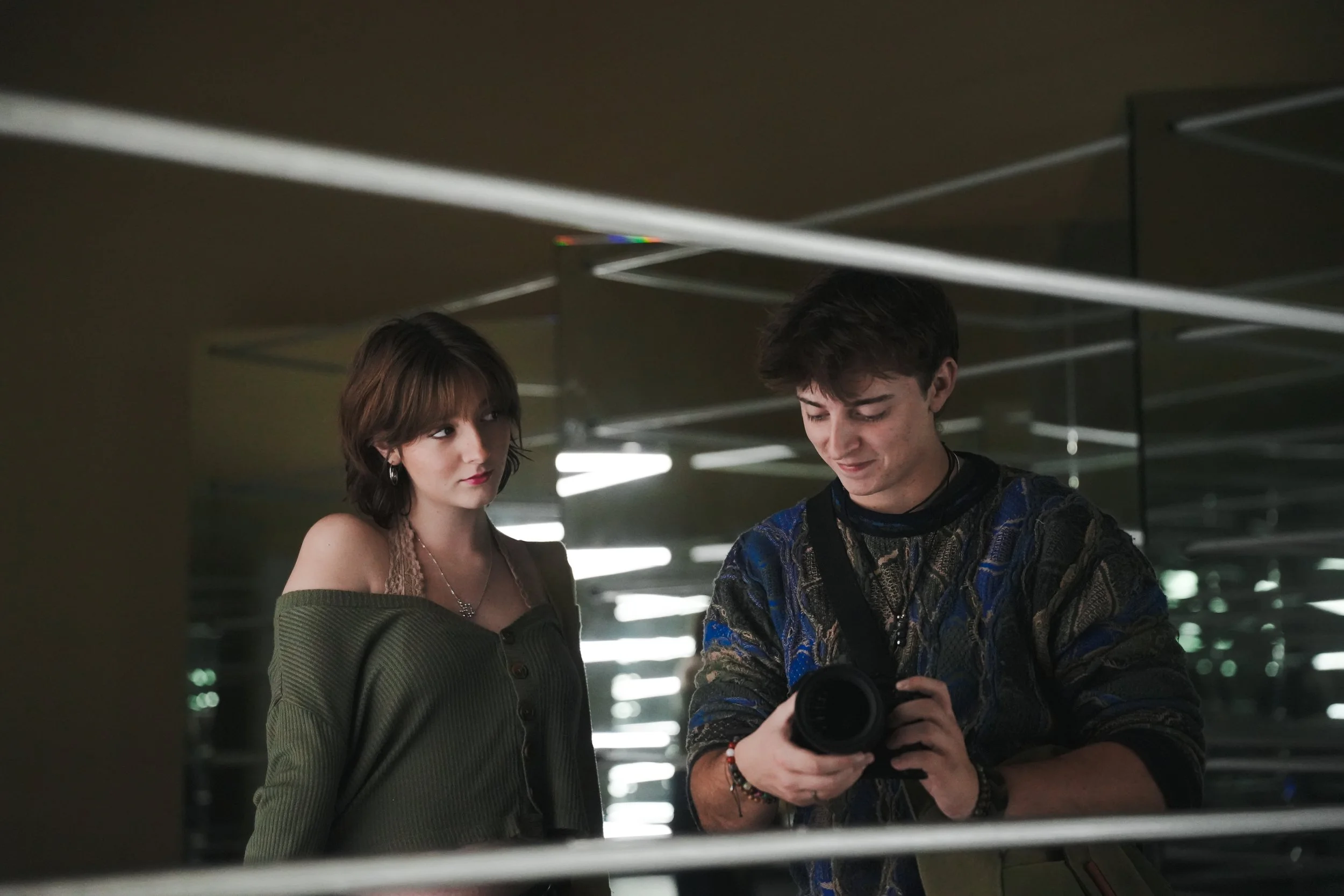 A young man and woman are looking at a camera held by the young man, in an indoor setting with mirrored walls and ceiling lights.