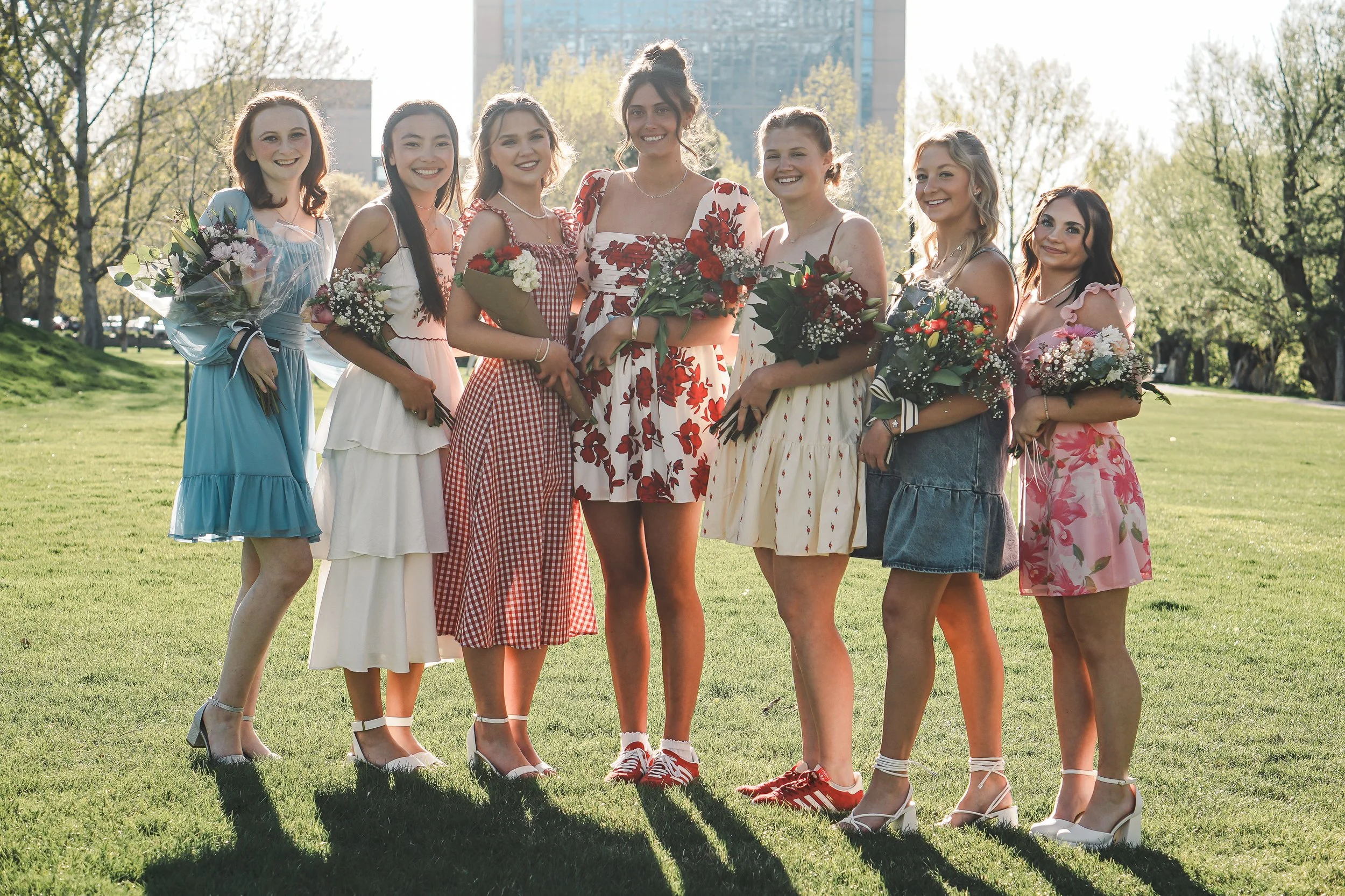 Eight young women standing outdoors on a sunny day in a grassy park, dressed in colorful summer dresses, holding bouquets of flowers, and smiling at the camera.