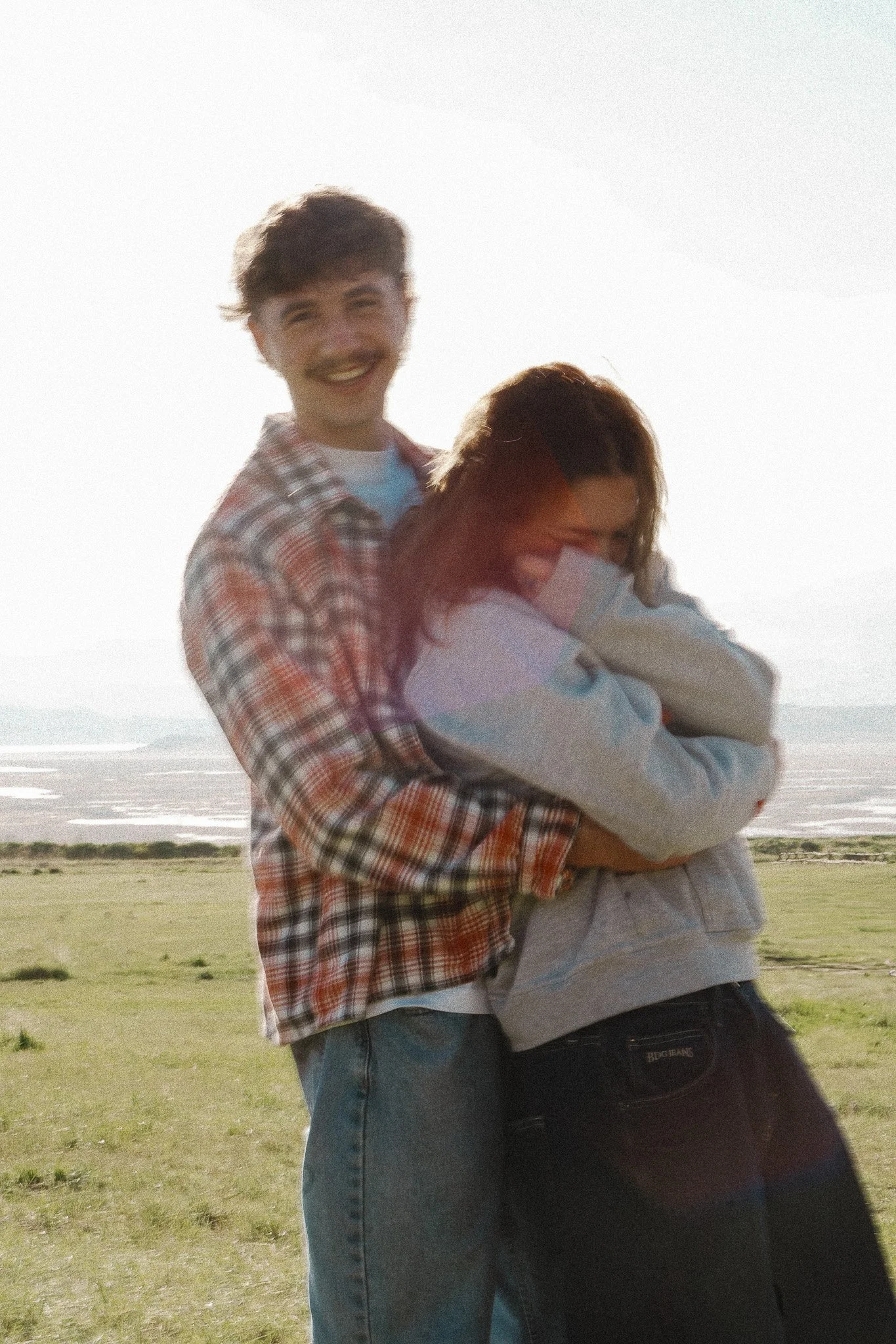 Young man and woman laughing and embracing outdoors on a grassy field near water, with the sun setting or rising in the background.